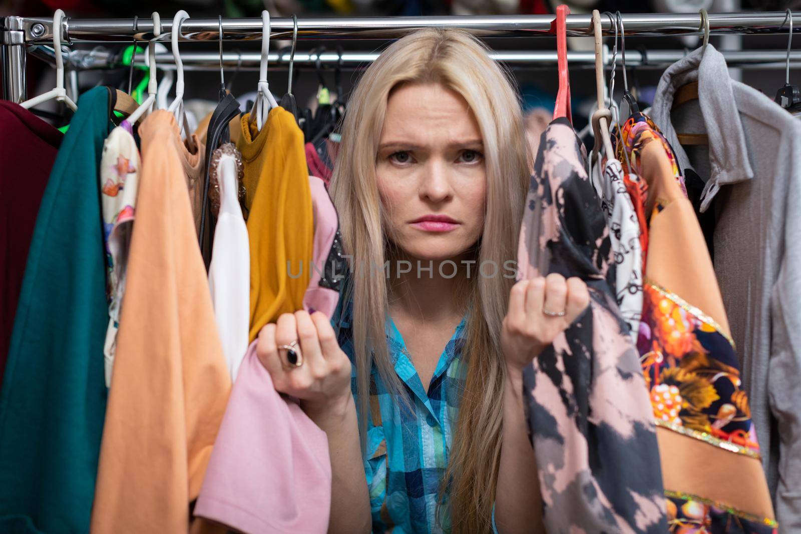 A blonde with a sour face. Shopping in a clothing mall. Hangers of clothes. She clenches her fists in anger and upset.