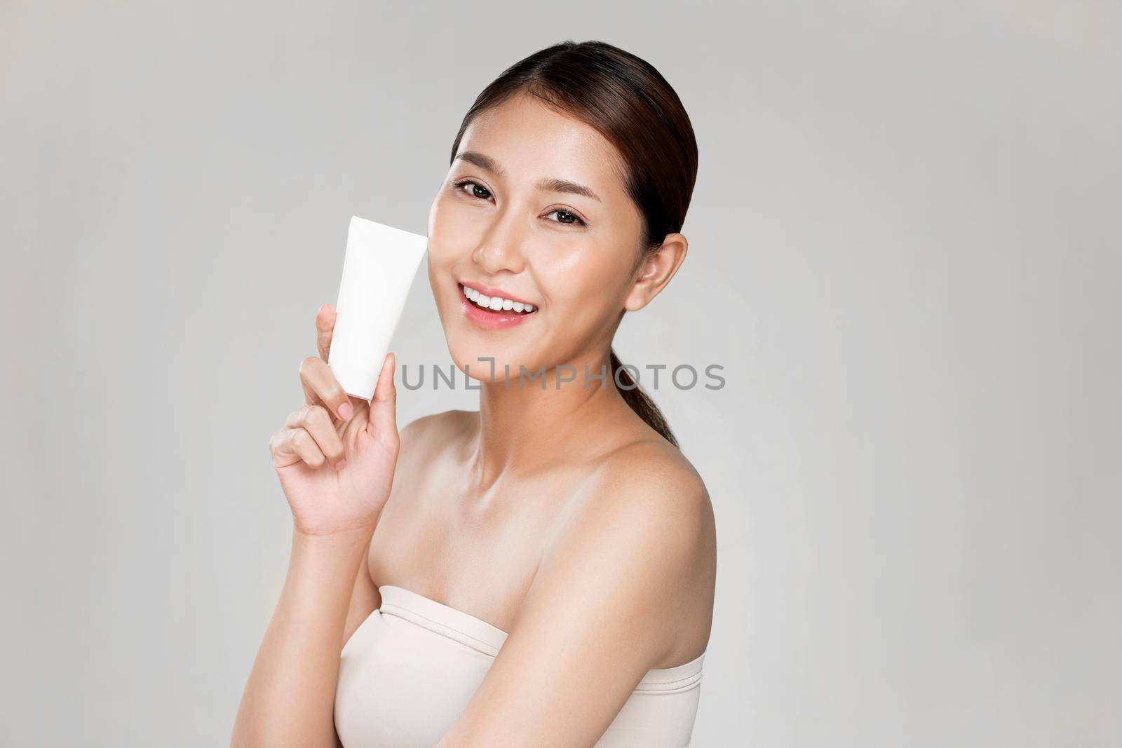 Ardent woman smiling holding mockup product for advertising text place, light grey background. Concept of healthcare for skin, beauty care product for advertising.