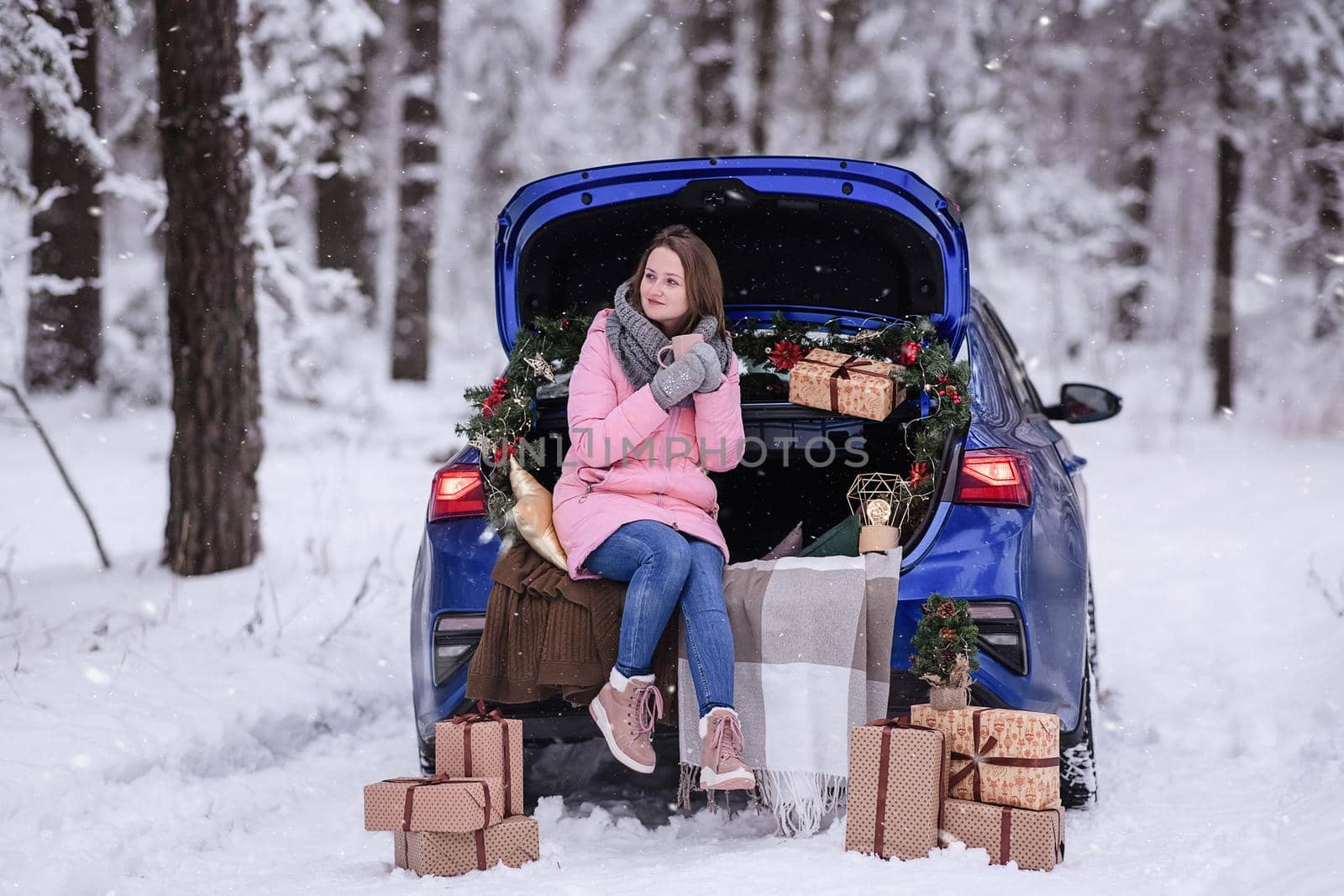 A woman in winter clothes drinks a hot drink sitting in a car decorated in a New Year's way. Traveling by car through the forest. A trip before Christmas.