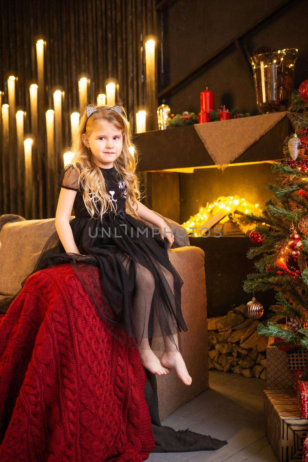 A girl in a festive outfit on the sofa with her dog next to a Christmas tree decorated with garlands, balloons and Christmas toys. The concept of winter holidays is Christmas and New Year holidays. Magical festive atmosphere.