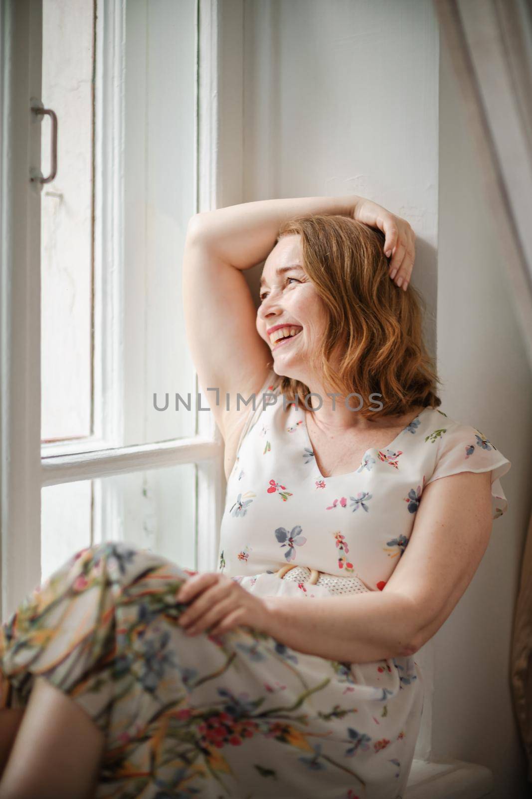 A middle-aged woman in a cream dress sits mysteriously and looks out the window on the windowsill. Green trees outside