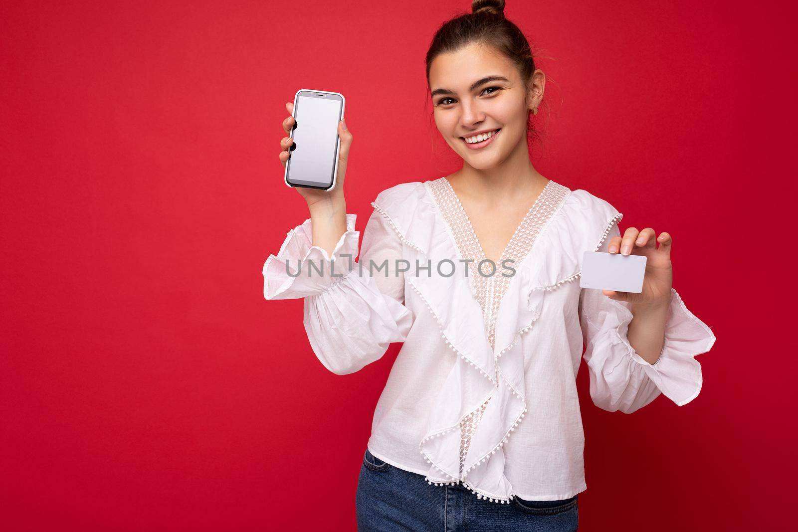 Portrait of positive cheerful fashionable woman in formalwear holding and showing mobile phone with empty screen for mock up and credit card with cut out looking at camera isolated on red background with copy space by TRMK