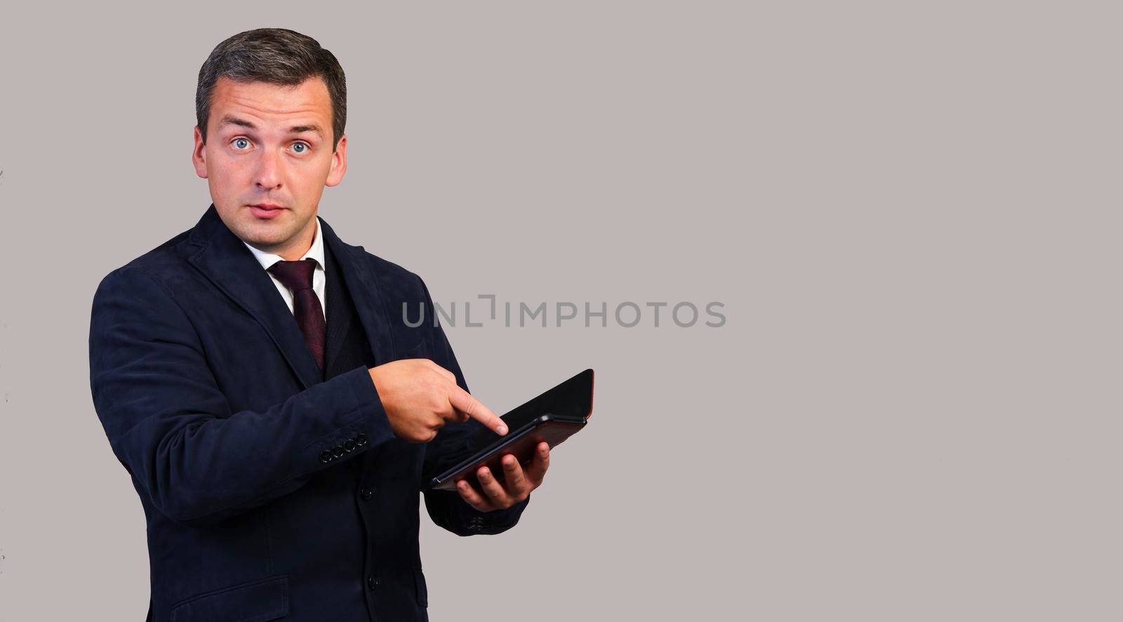 A handsome man in a blue jacket with a tie with an e-book in his hands points to her with his index finger on a yellow background.