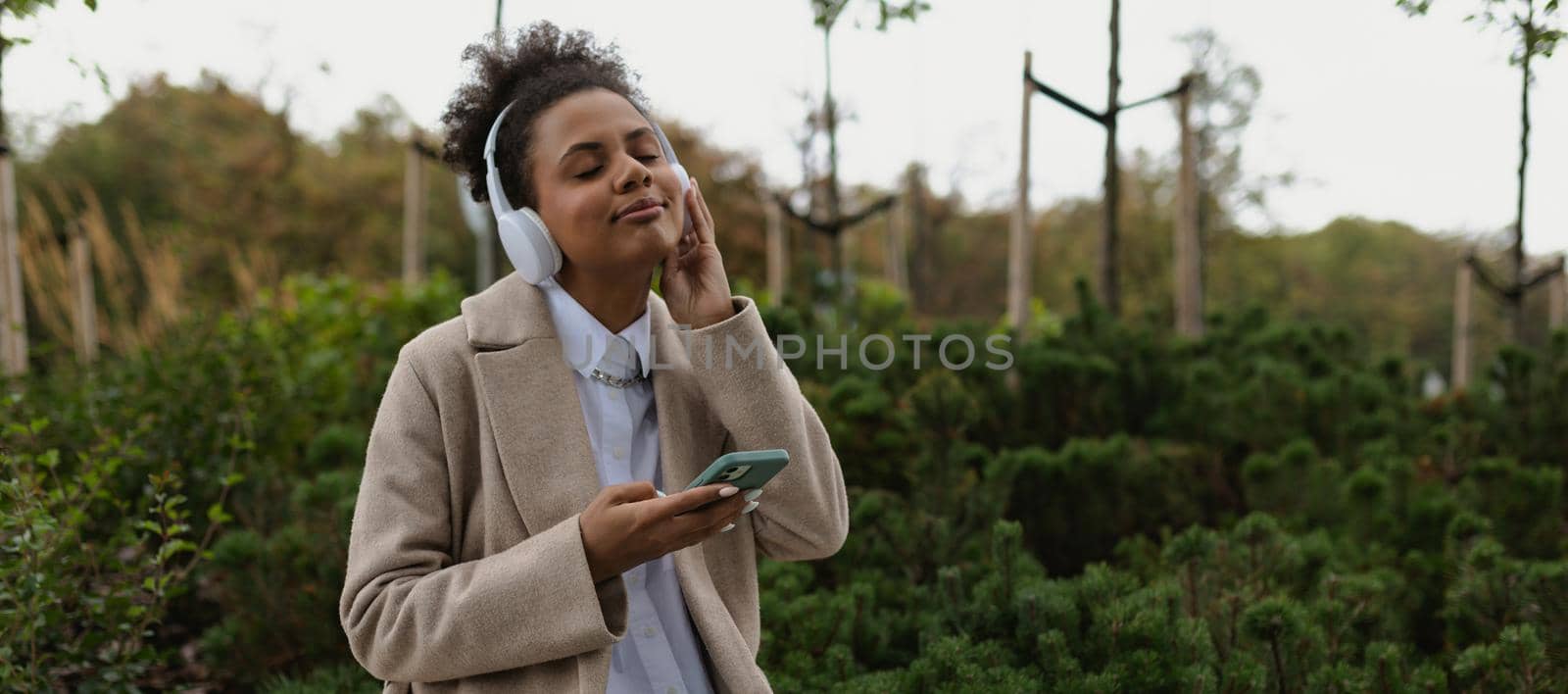 a young african american woman in a beige coat listens to music in headphones with her eyes closed against the backdrop of a city park by TRMK