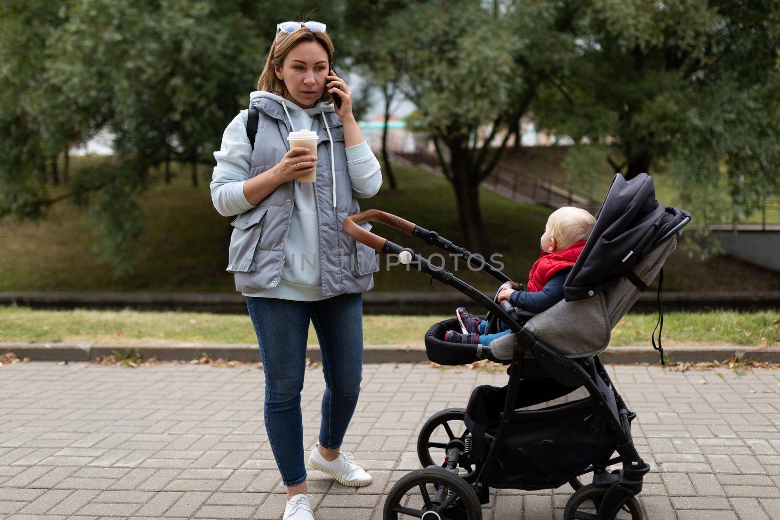 young woman while walking with her baby and pram talking on a mobile phone and drinking coffee in a city park.