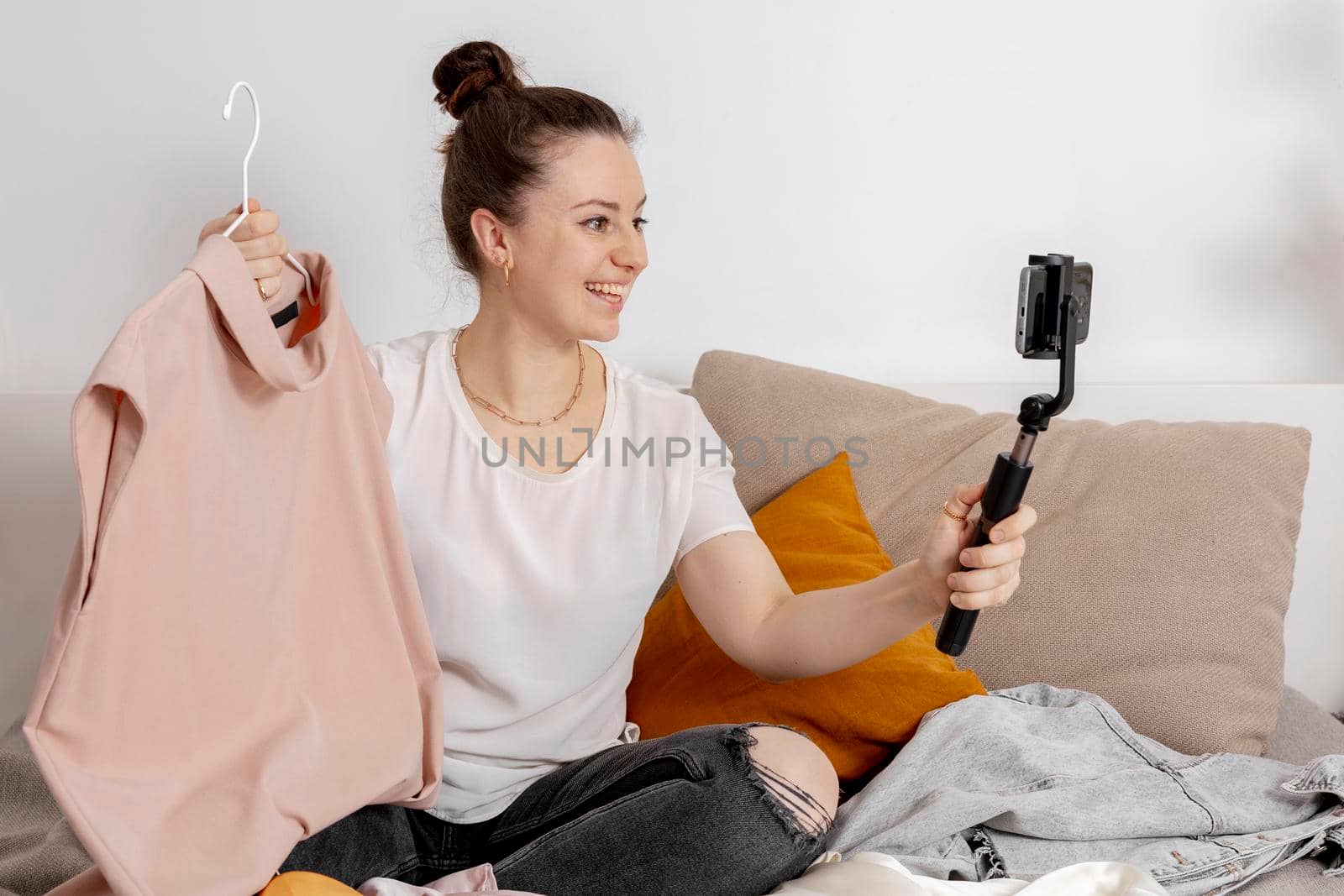 Young woman sitting on the bed at home and taking photo of her old clothes to sell them online. Selling on website, e-commerce. Reuse, second-hand concept. Conscious consumer, sustainable lifestyle