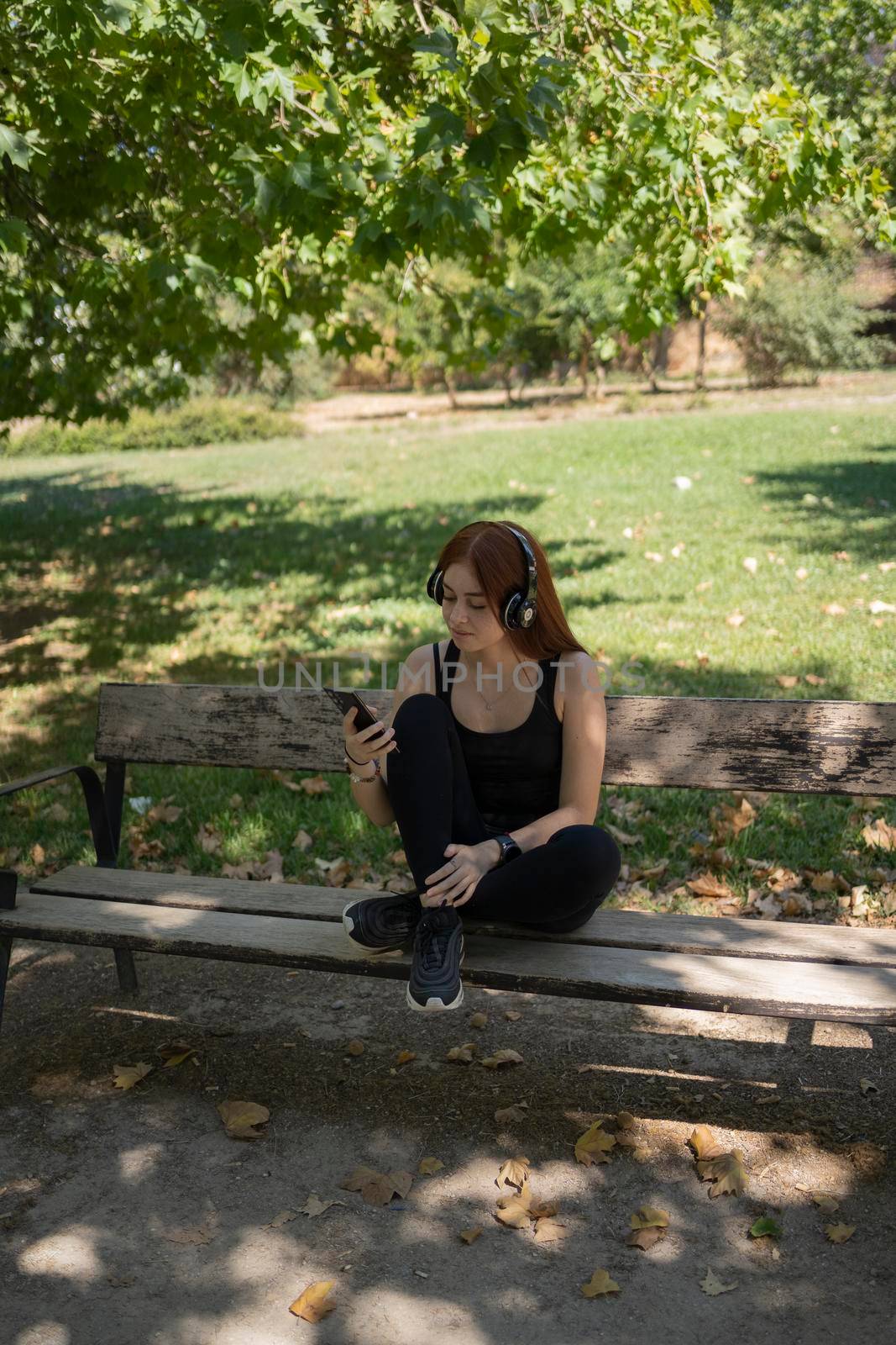 young redhead girl, looking at the smartphone, with music headphones sitting on a park bench