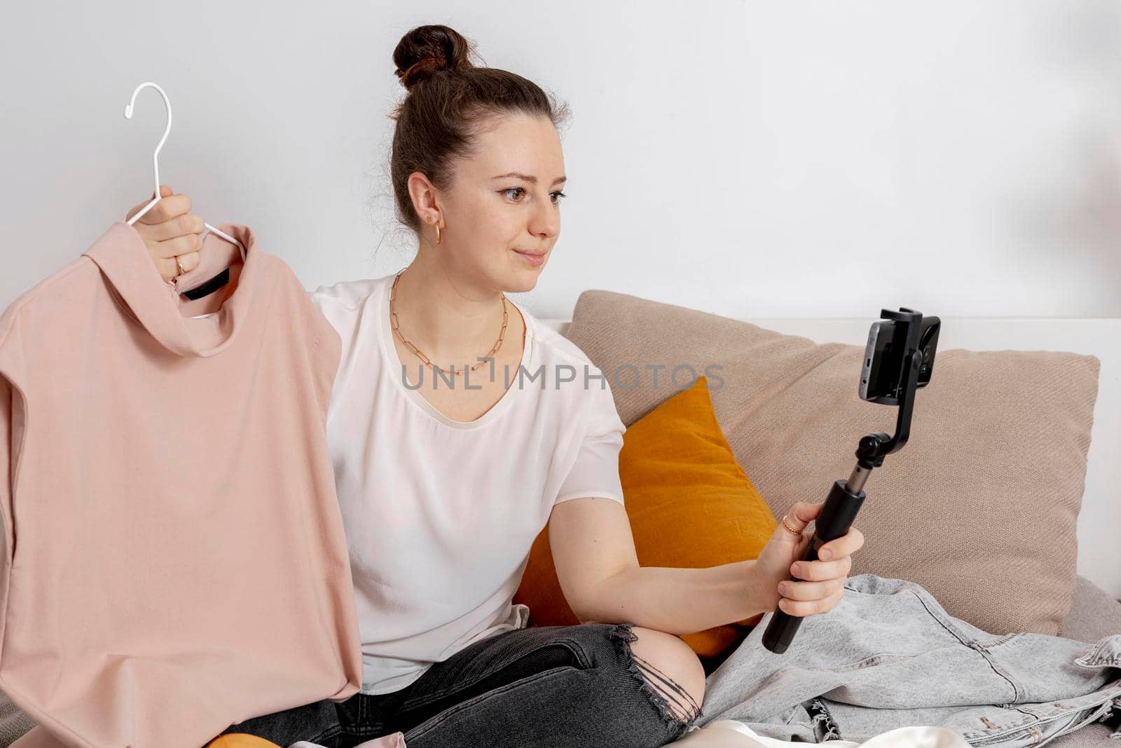 Young woman sitting on the bed at home and taking photo of her old clothes to sell them online. Selling on website, e-commerce. Reuse, second-hand concept. Conscious consumer, sustainable lifestyle