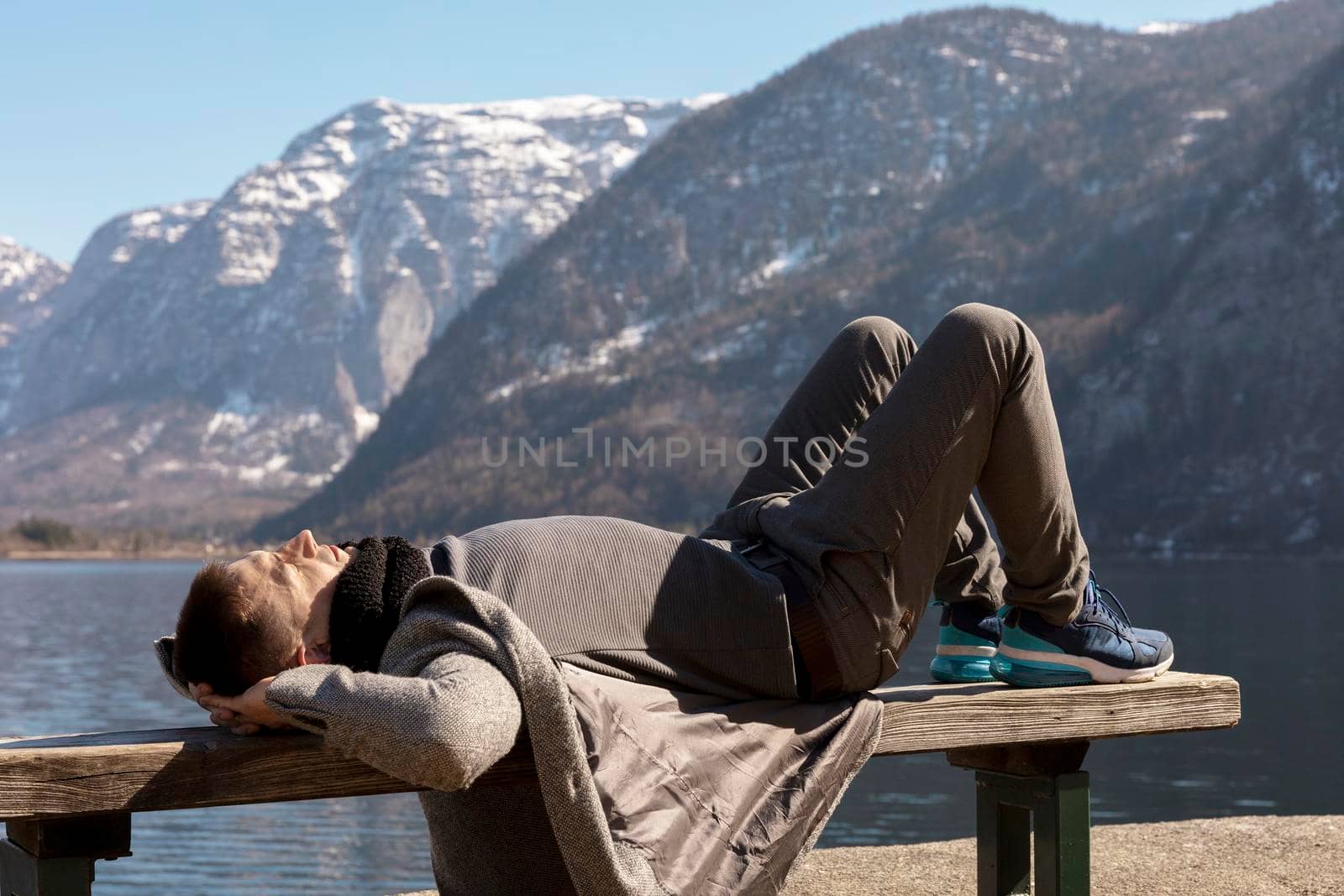 Young man lying outdoors on bench and enjoying mountains, snow, good weather, blue sky, sun. Beautiful landscape. Time with yourself, dreaming, relaxation, mental health. Tourism, holiday, travel