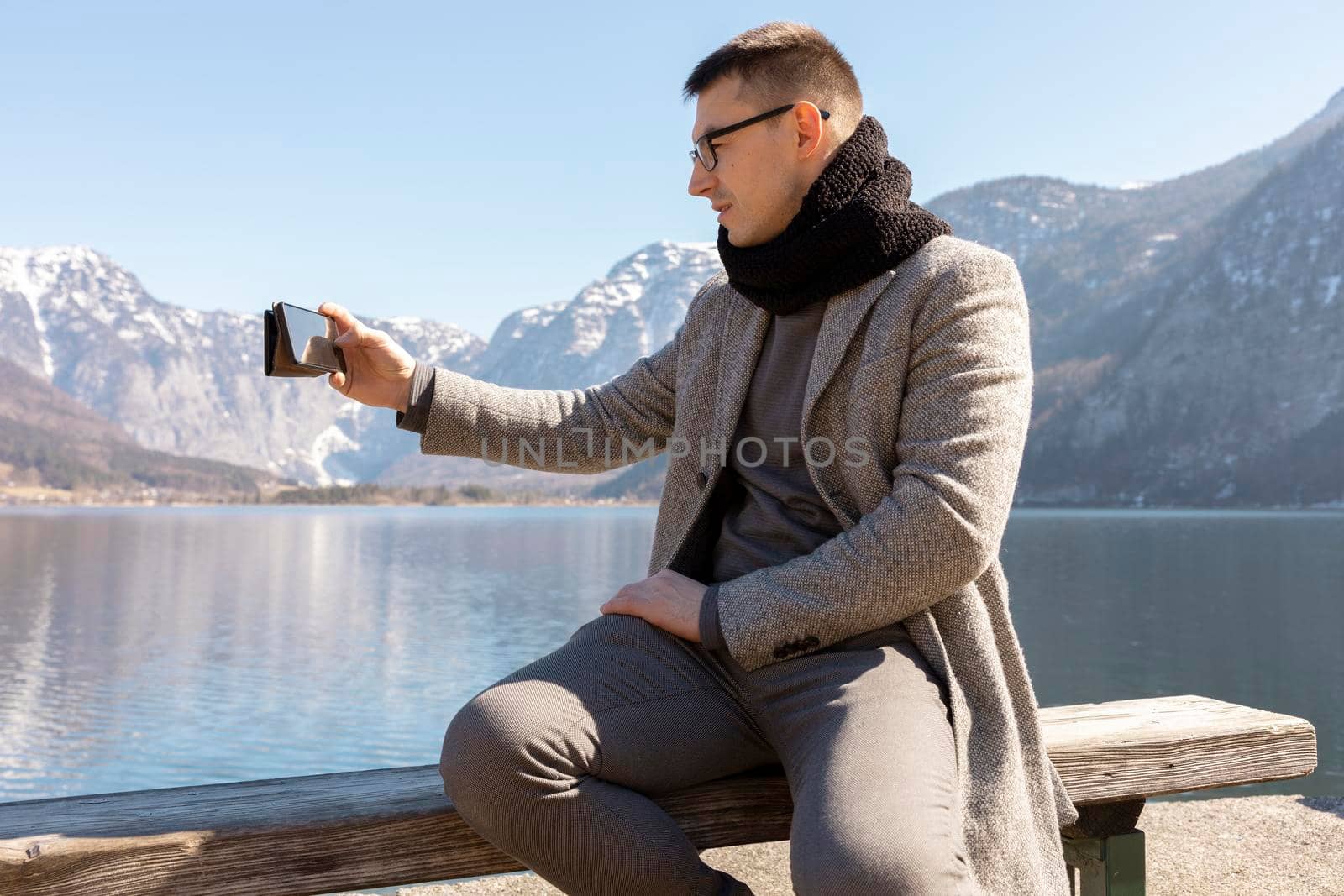 Young adult man sitting outdoors on bench and making selfie with his smartphone, enjoying mountains, lake, good weather, blue sky, sun. Beautiful, amazing landscape. Holiday, vacation, travel time. by creativebird