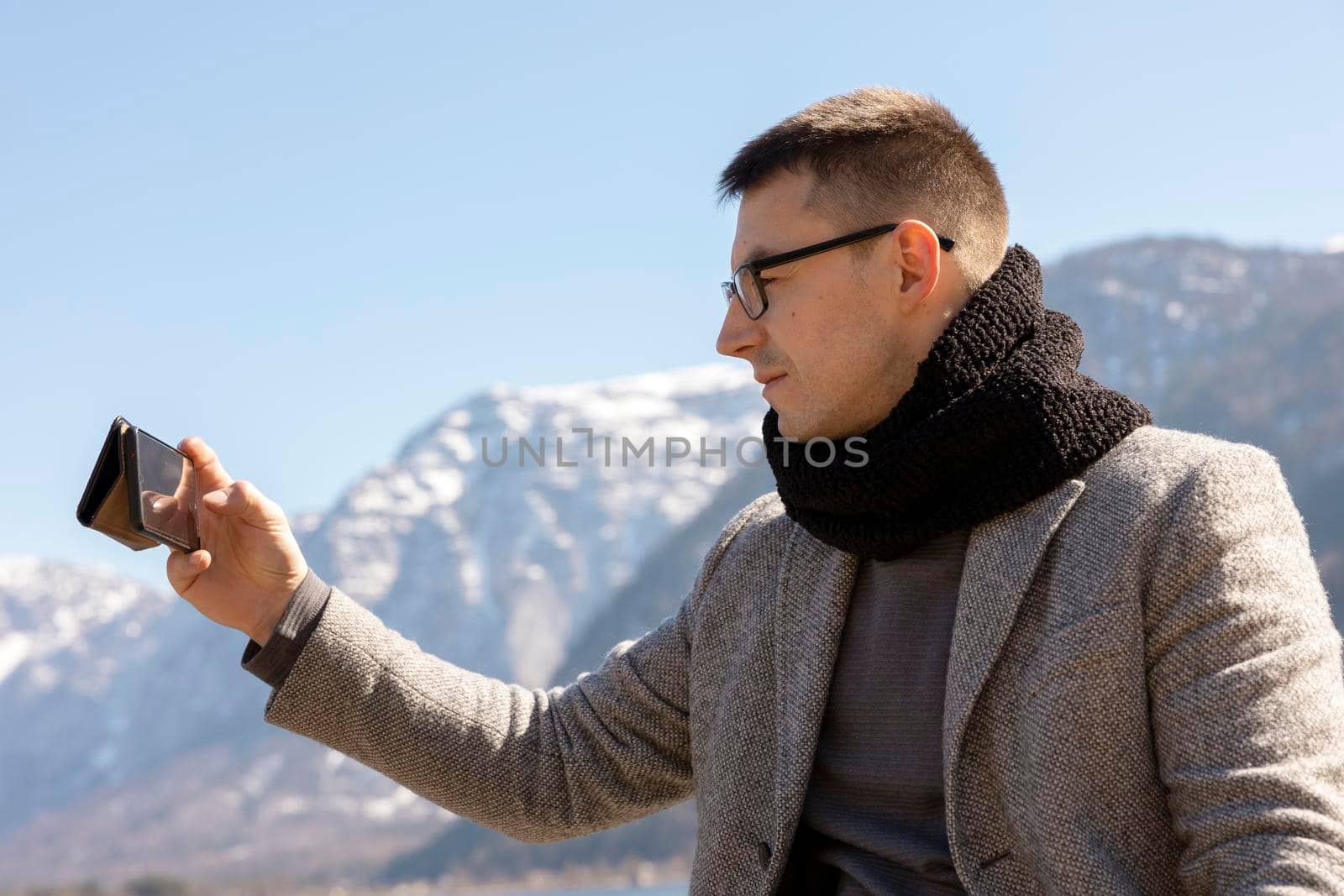 Young adult man sitting outdoors on bench and making selfie with his smartphone, enjoying mountains, lake, good weather, blue sky, sun. Beautiful, amazing landscape. Holiday, vacation, travel time