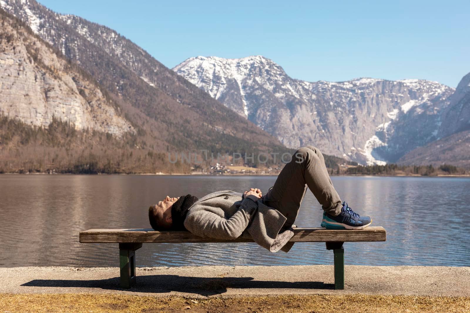 Young man lying outdoors on bench and enjoying mountains, snow, good weather, blue sky, sun. Beautiful landscape. Time with yourself, dreaming, relaxation, mental health. Tourism, holiday, travel