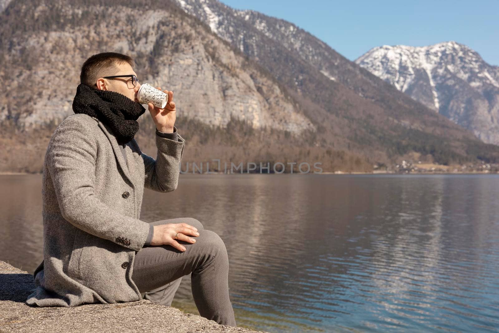 Young adult man sitting outdoors, drinking coffee and enjoying mountains, lake, good weather, blue sky and sun. Beautiful landscape. Time with yourself, dreaming, relaxation, mental health. Holiday. by creativebird