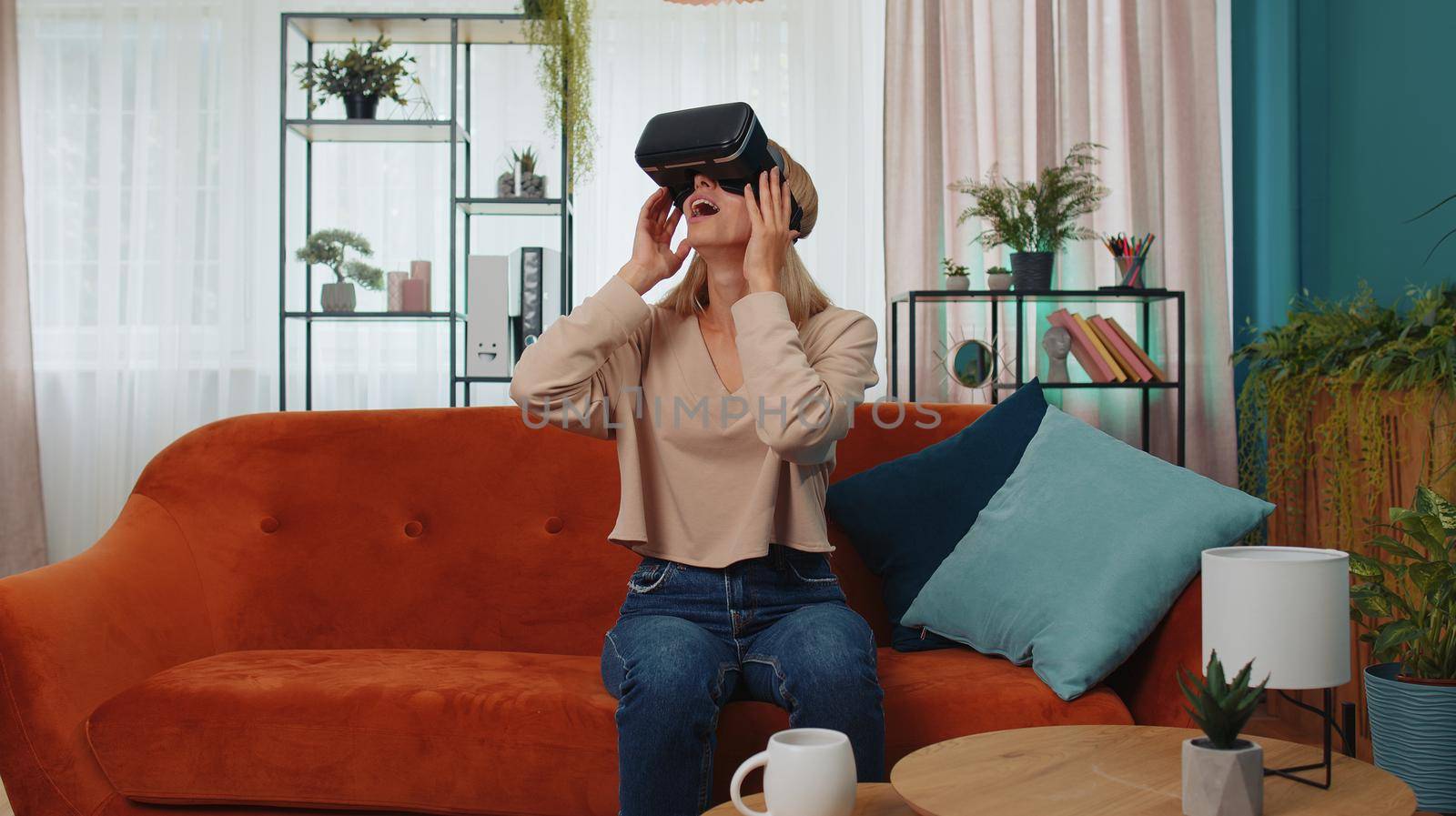 Young woman use VR headset glasses at home, enjoying 3D 360 video concept, entertaining, in positive mood, moving hands in air, sitting on sofa in living room. Virtual reality, modern gadget device