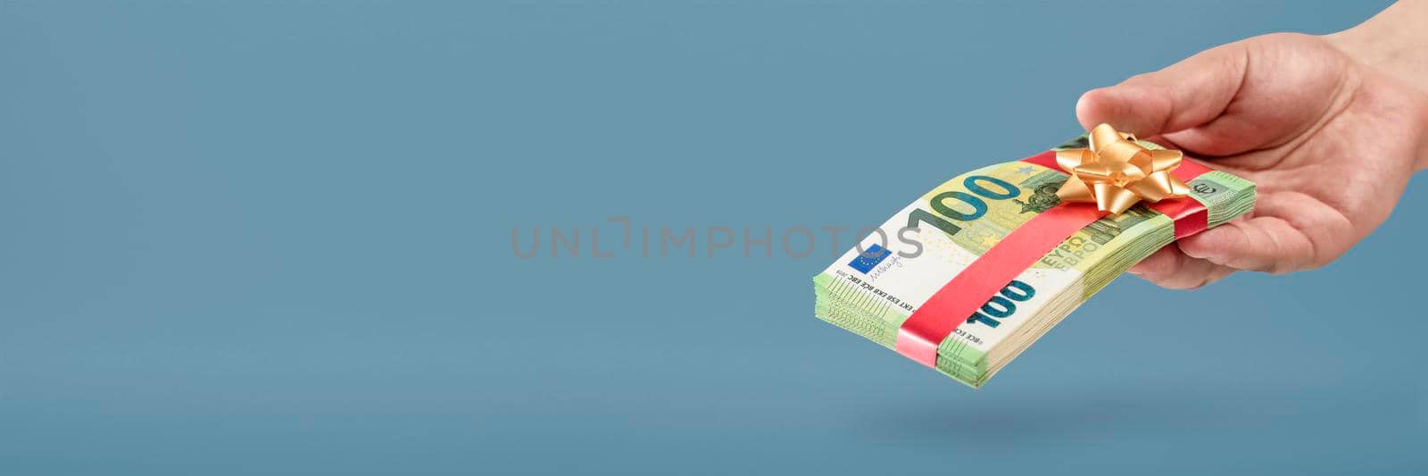 Gift of money. A man holds a stack of banknotes in his hands as a symbol or concept of a gift. 100 euro banknotes tied with a red ribbon with a bow on a blue background