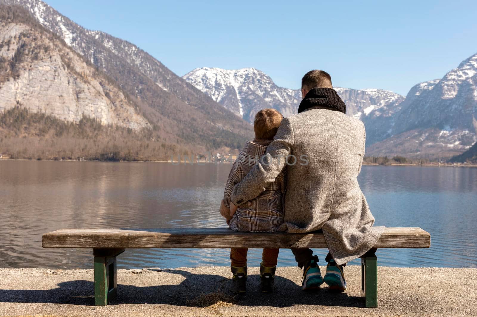 Father and son sitting together outdoors on the bench and enjoying mountains, snow, good weather, blue sky. Little boy and his father spending time together. Family time. Beautiful winter landscape