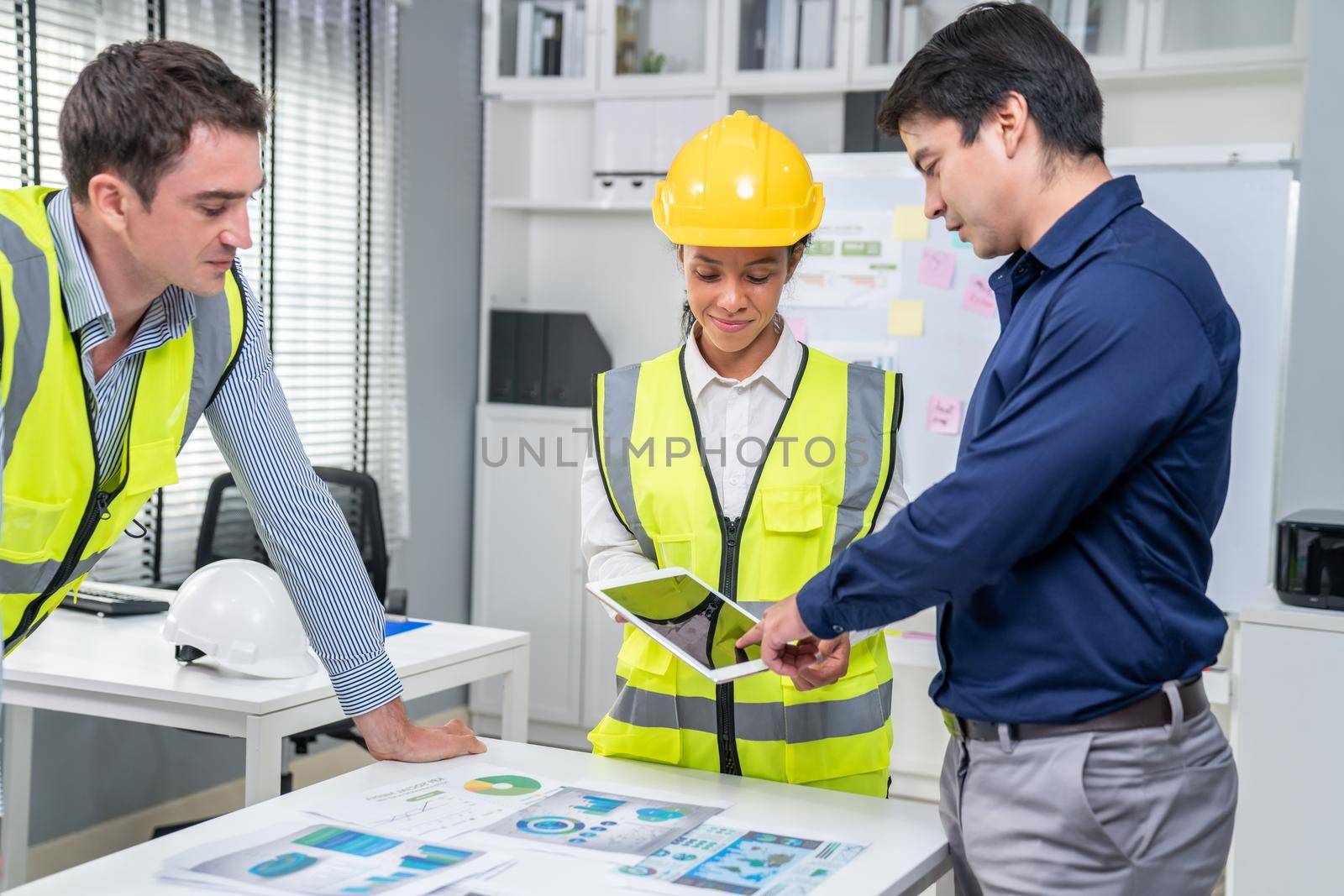 A team of competent engineers wearing safety equipment is working on blueprints with a tablet while also discussing with the investor in the office.