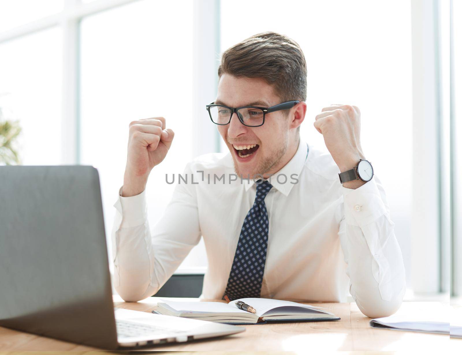Happy young businessman raising hands in front of laptop at office desk by Prosto