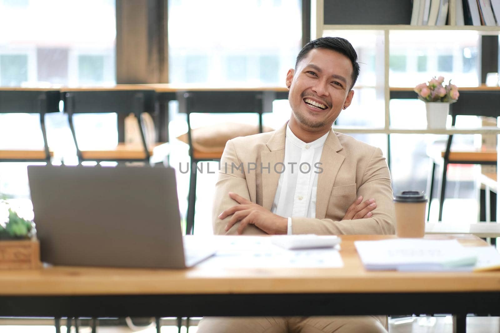 Portrait of successful asian businessman standing with arms crossed at desk smiling to camera.