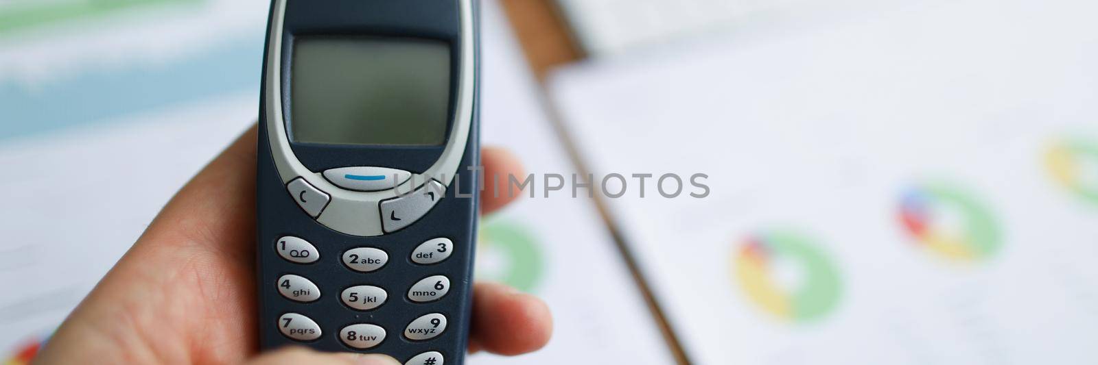 In the hands of an old model of a mobile phone against the background of reports on the table, close-up, blurry. Corporate cell phone