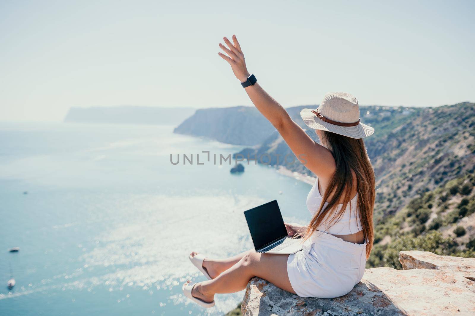 Successful business woman in yellow hat working on laptop by the sea. Pretty lady typing on computer at summer day outdoors. Freelance, travel and holidays concept.