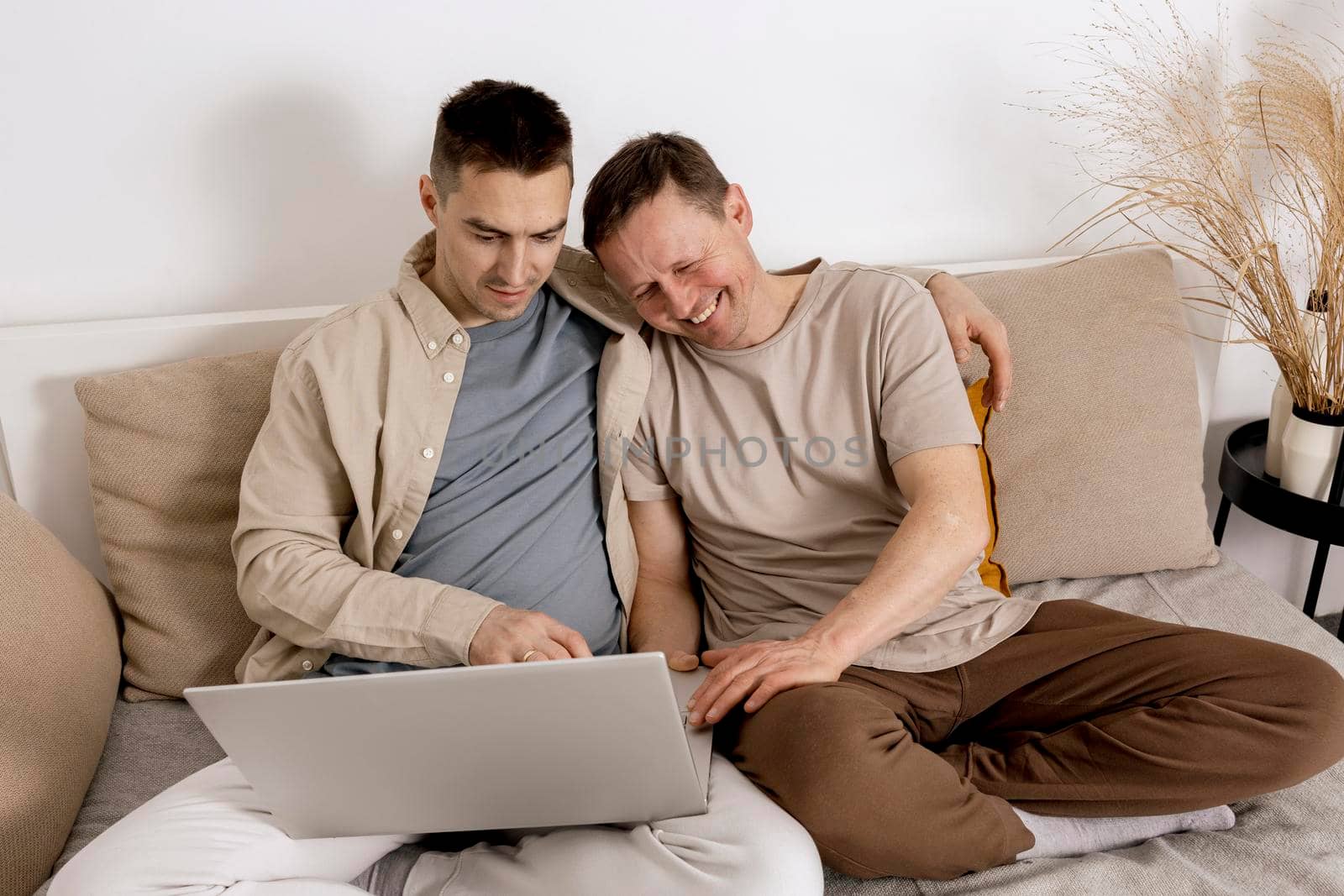 Happy gay couple with casual clothes spending time together at home and watching movie on the laptop. Two caucasian men relaxing. Homosexual relationships and alternative love. Cosy interior