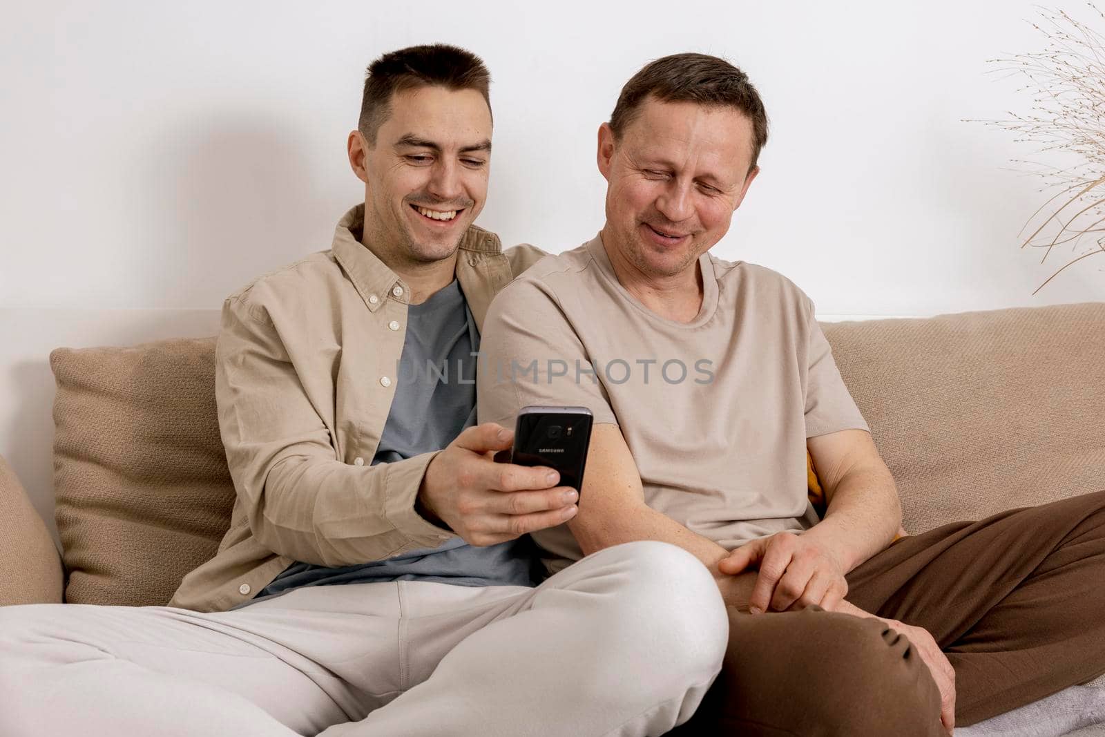 Happy gay couple with casual clothes spending time together at home and watching something on smartphone, or having video call with friends. Homosexual relationships and alternative love