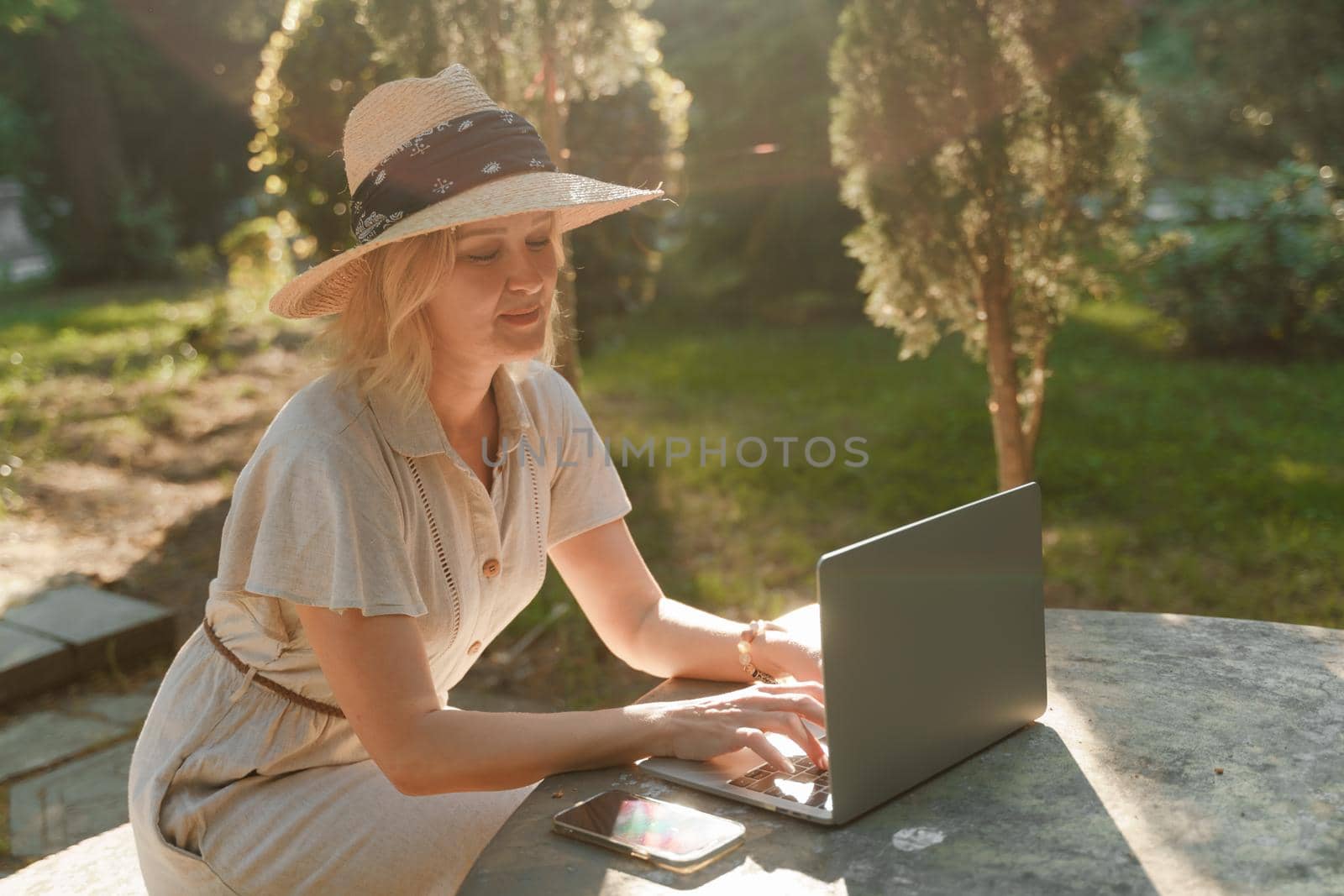 a beautiful mature woman in hat works on a computer at a white table in nature and spends her day productively.
