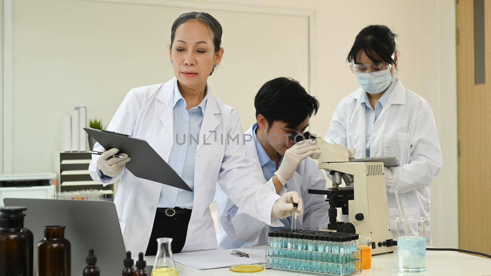 Senior female scientist analyzing and summarising information for scientific report and laboratory assistant working in background by prathanchorruangsak