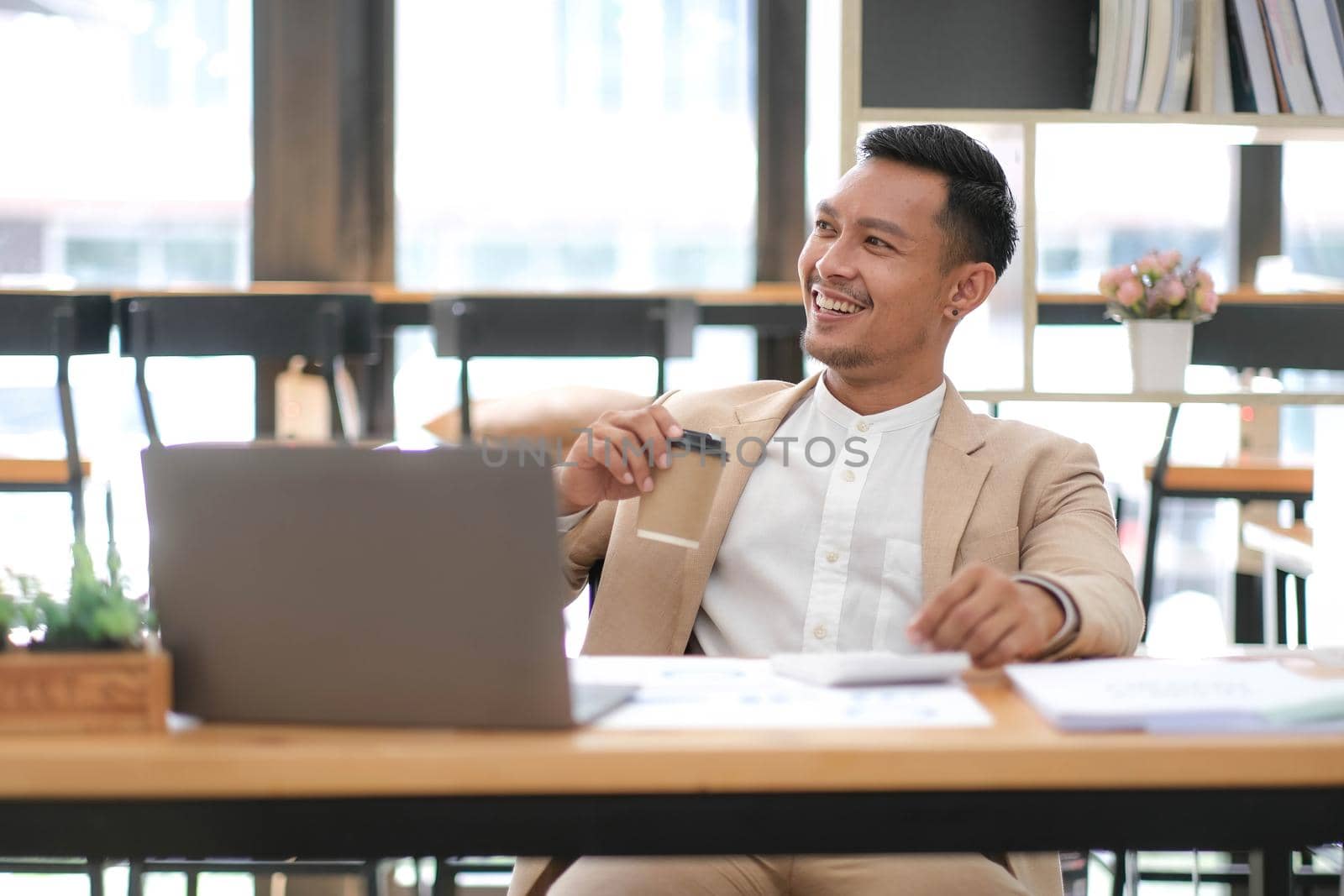 Young Asian businessman in suit and holding hot coffee cup and looking on laptop computer in office. Man in suit using laptop in well-lit workplace. by wichayada