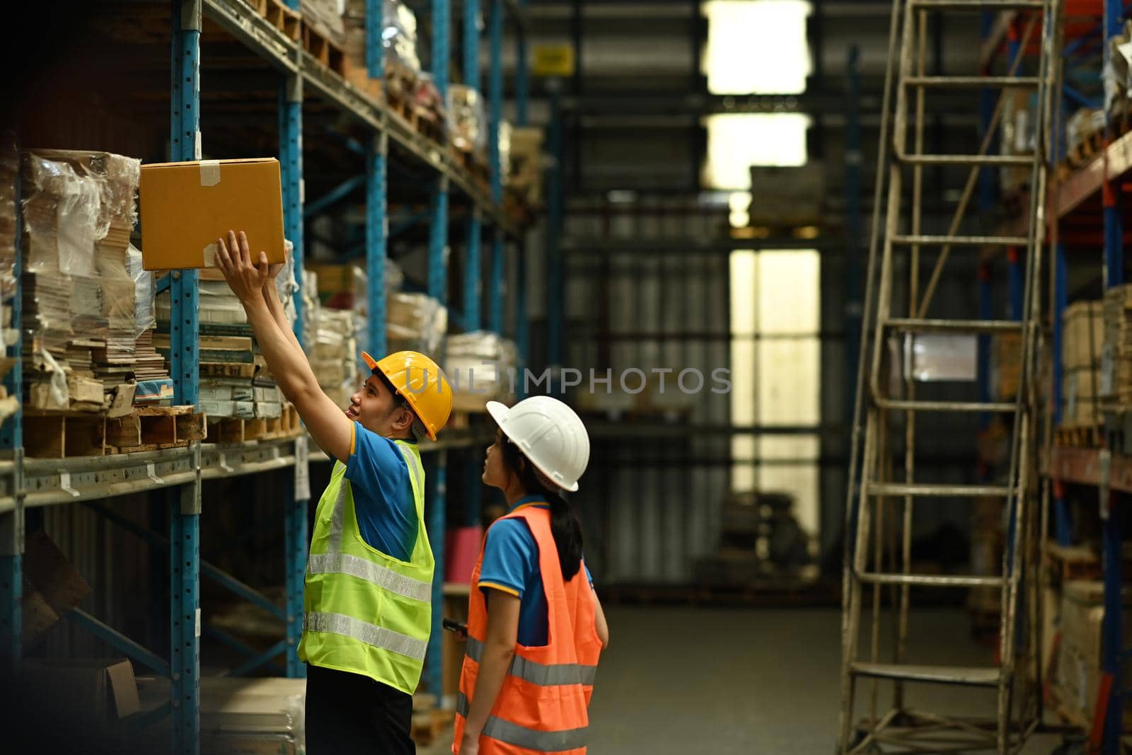 Two young colleagues wearing hardhat and reflective jacket checking stock on the shelves in a large warehouse.