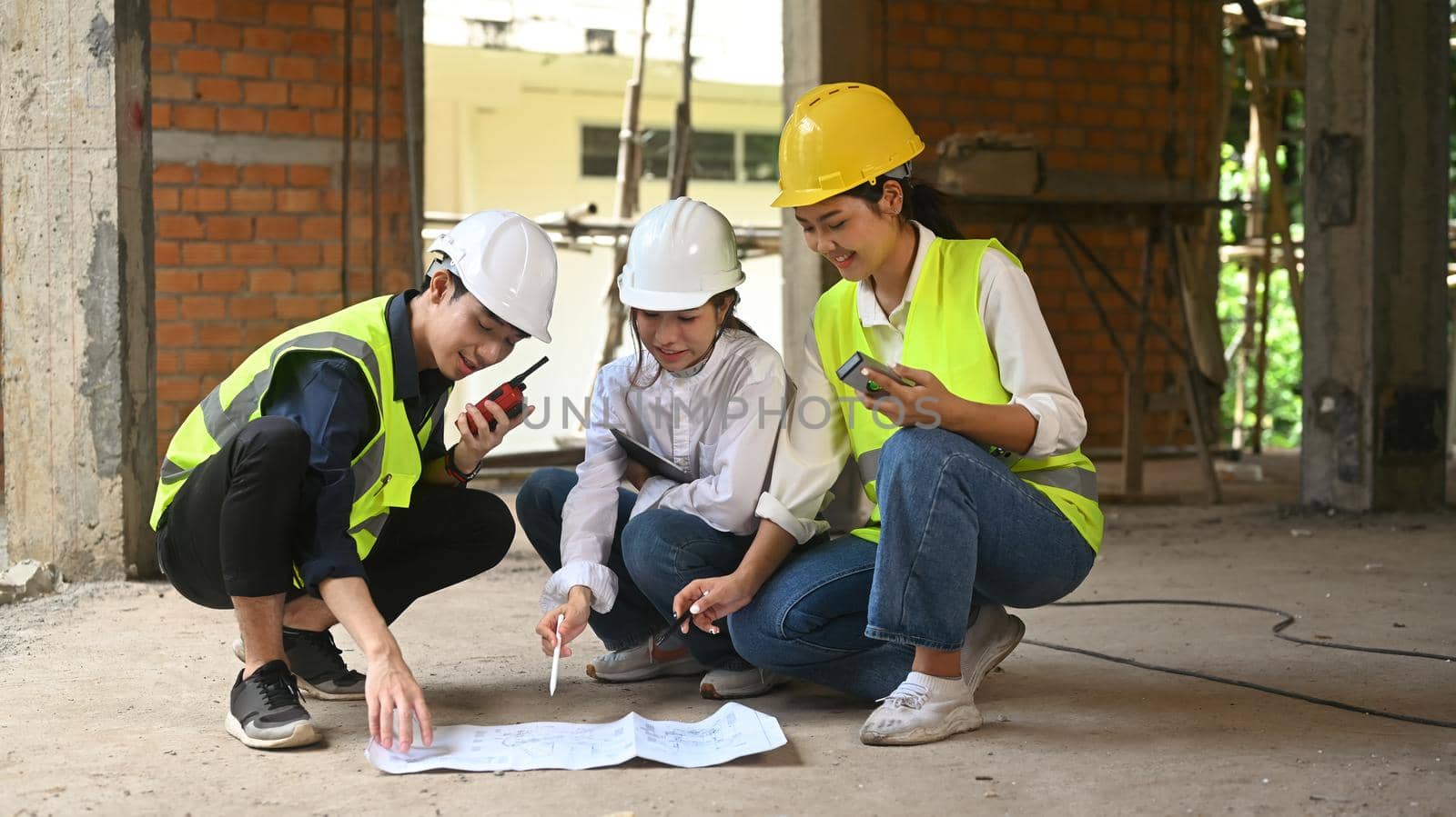 Civil Engineer manager and Architect team in safety helmet are discussing plan details together while sitting under new building.