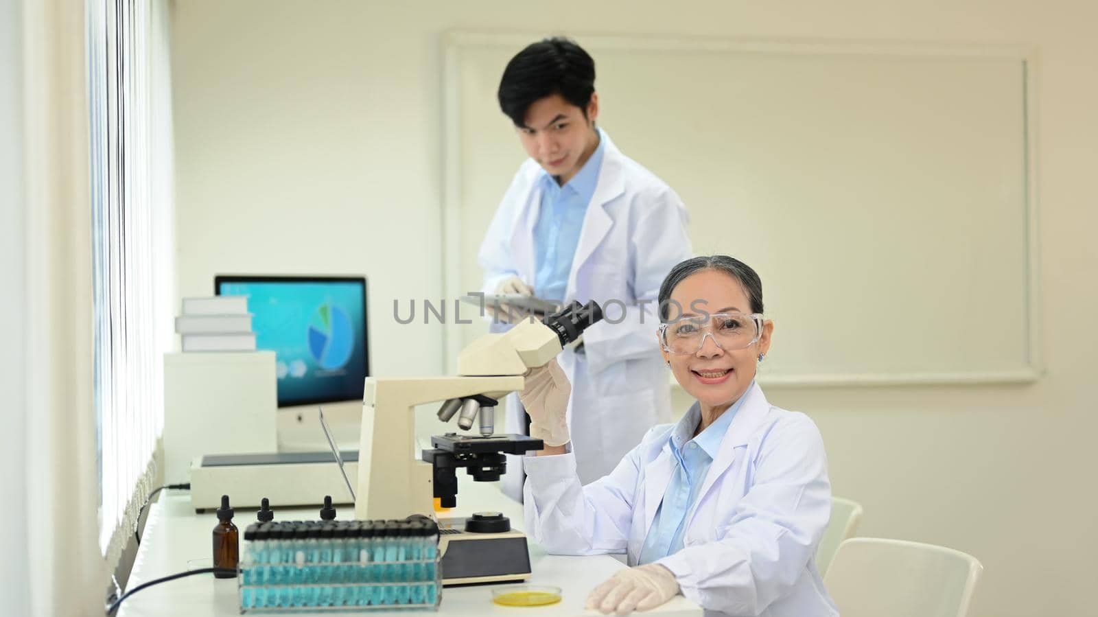 Experienced senior woman scientist in white coat sitting front of microscope and smiling to camera by prathanchorruangsak
