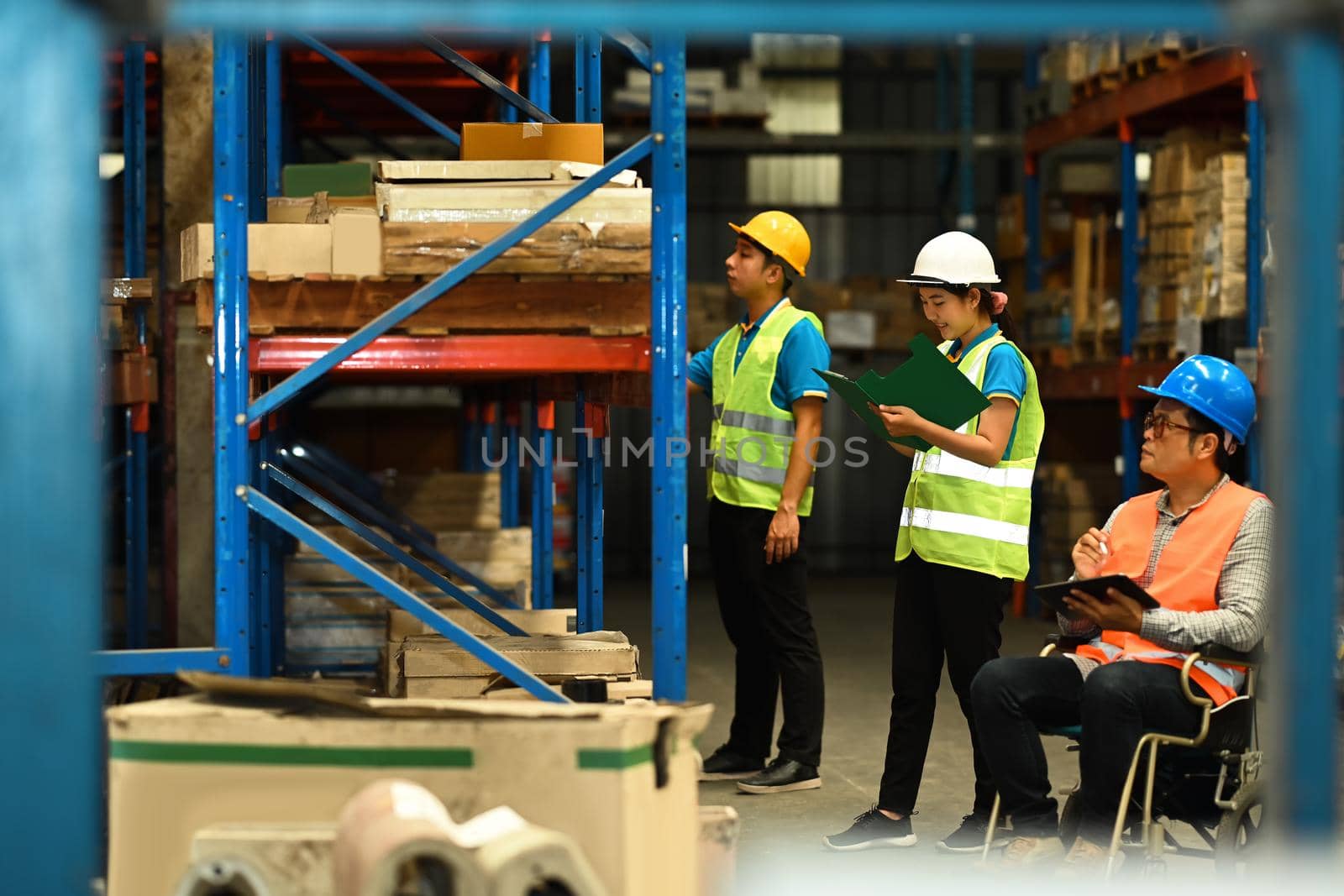 Male manager in wheelchair and young storehouse employees inspecting stock in a large warehouse by prathanchorruangsak