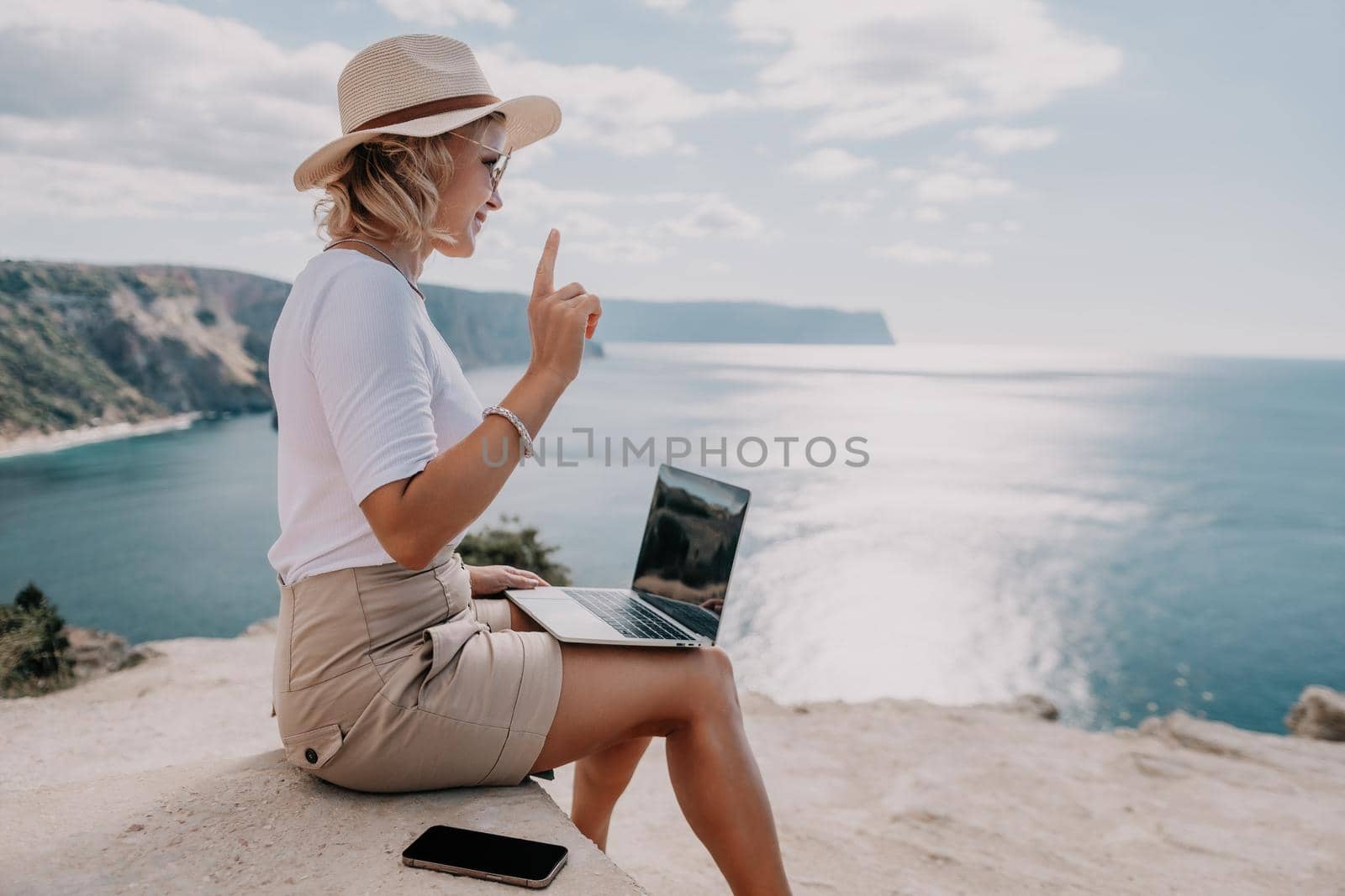 Successful business woman in yellow hat working on laptop by the sea. Pretty lady typing on computer at summer day outdoors. Freelance, travel and holidays concept.