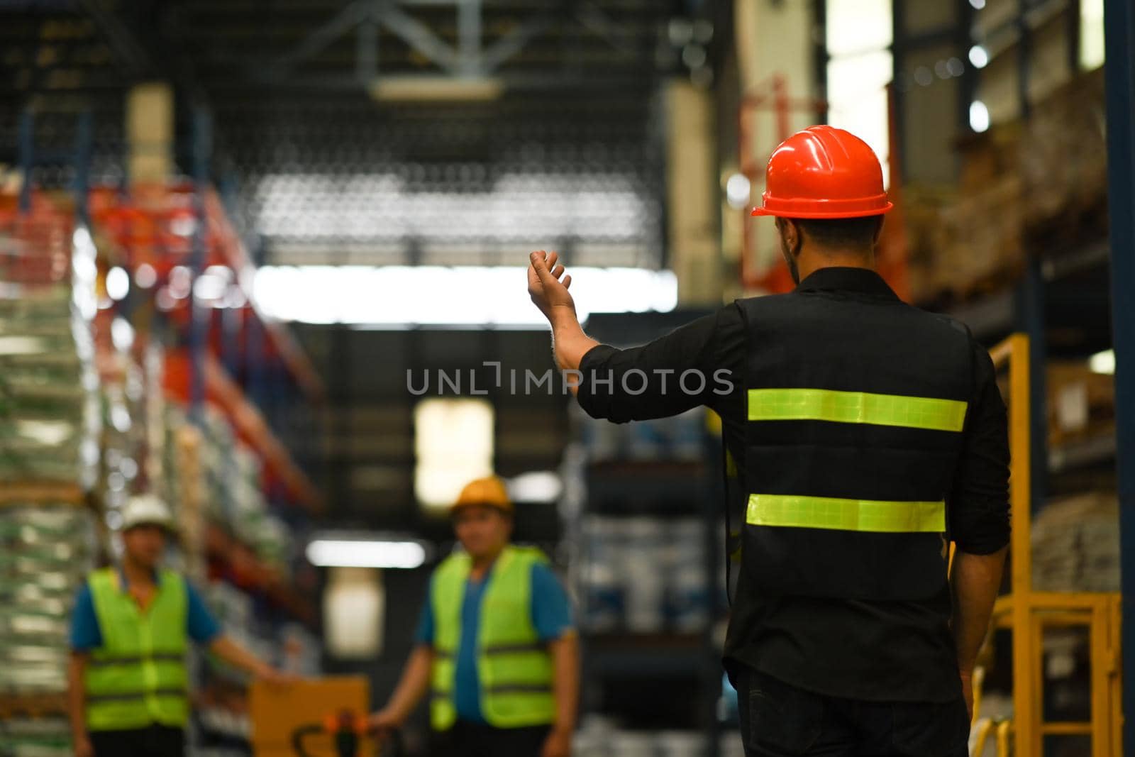 Back view of male manager wearing hardhat standing between retail full of shelves controlling work of industry logistics warehouse people by prathanchorruangsak