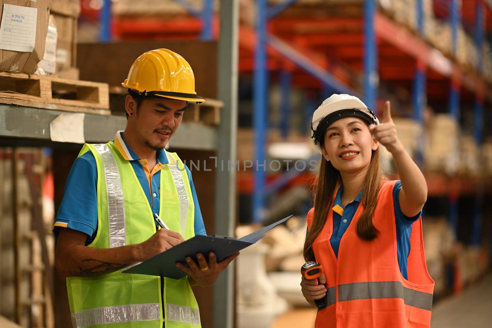 Warehouse workers wearing hardhats and reflective jackets using digital tablet, checking stock and order details in warehouse.