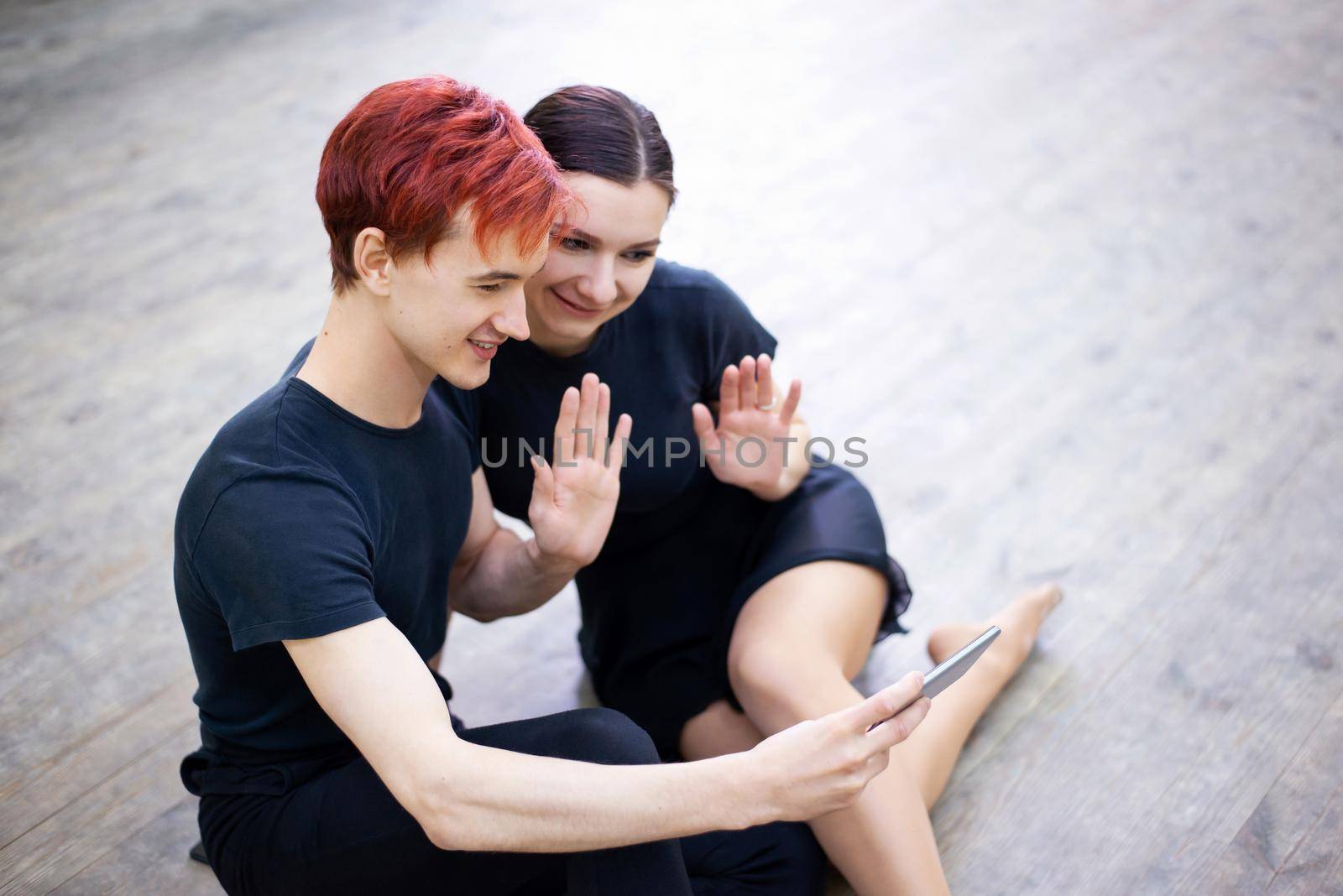 Two friends dancers in black clothes sitting on the floor and waving to other friend, while having a video chat by VitaliiPetrushenko