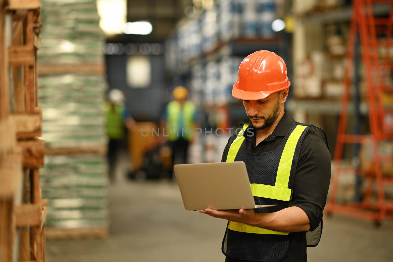 Warehouse manager wearing hardhats and reflective jackets using laptop for checking stock and order details in warehouse.