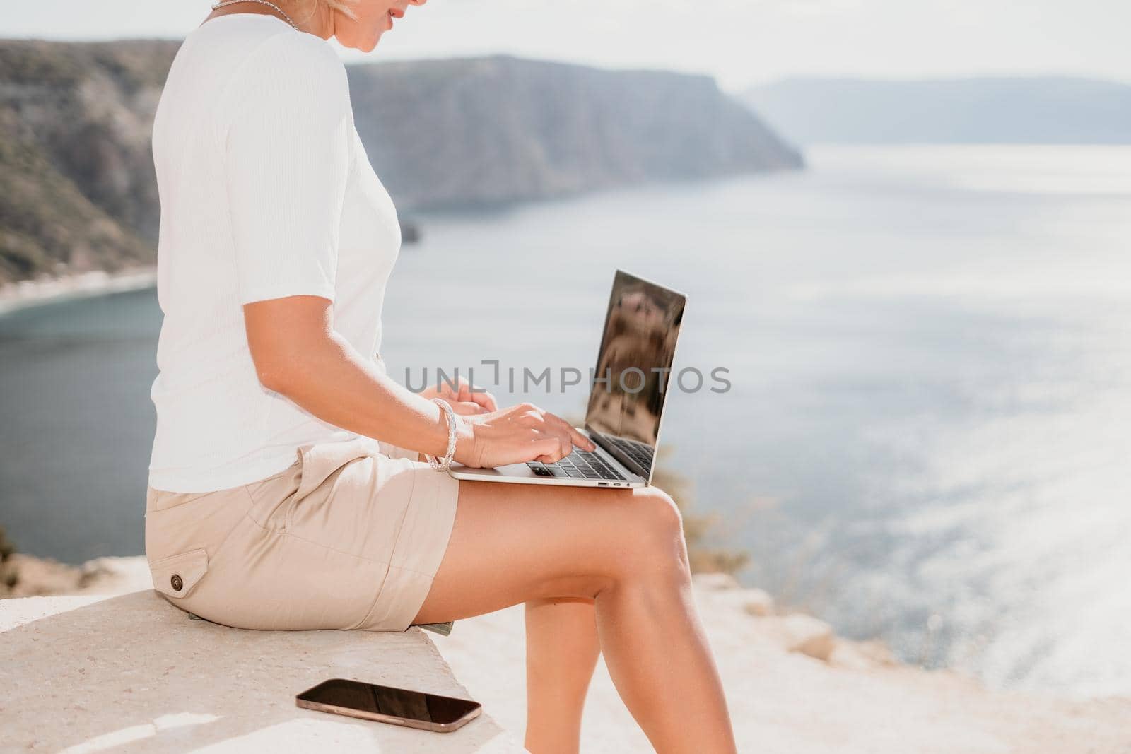 Successful business woman in yellow hat working on laptop by the sea. Pretty lady typing on computer at summer day outdoors. Freelance, travel and holidays concept.