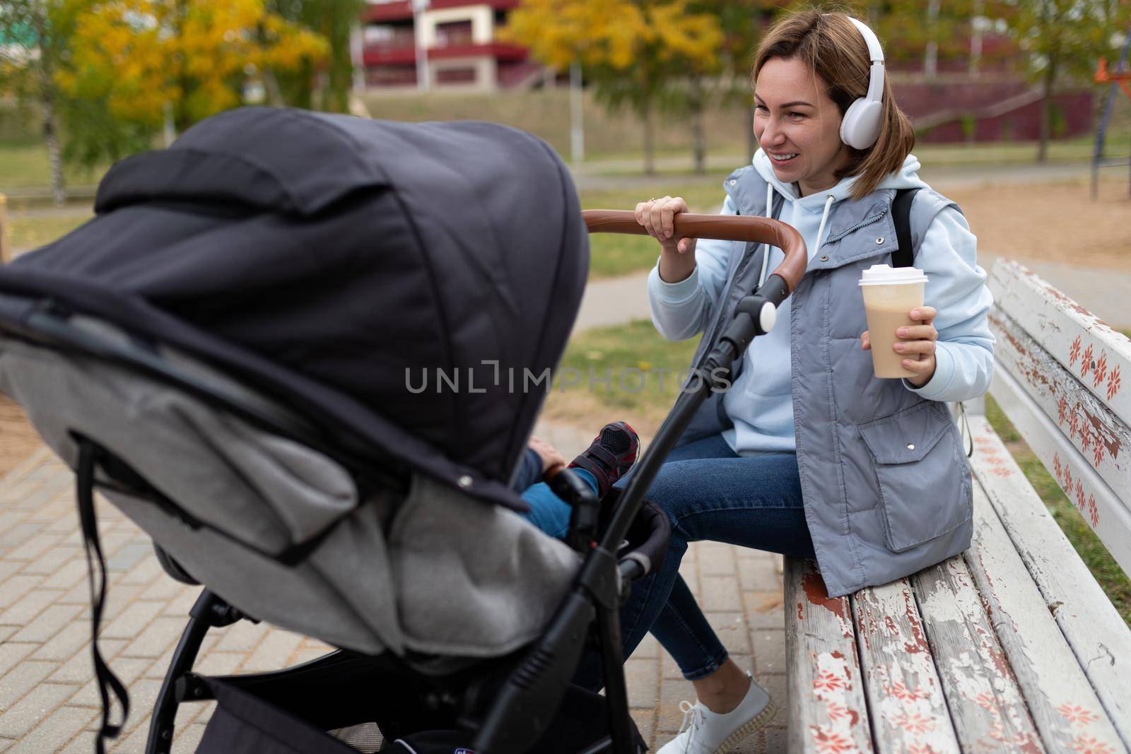 mother with a baby in a stroller sits on a bench and drinks coffee in headphones.