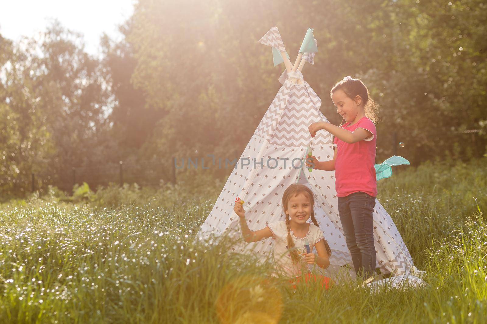 Two happy little kids are sitting on the grass and reading fairy tales near a tent in their home yard. Home camping vacation concept.