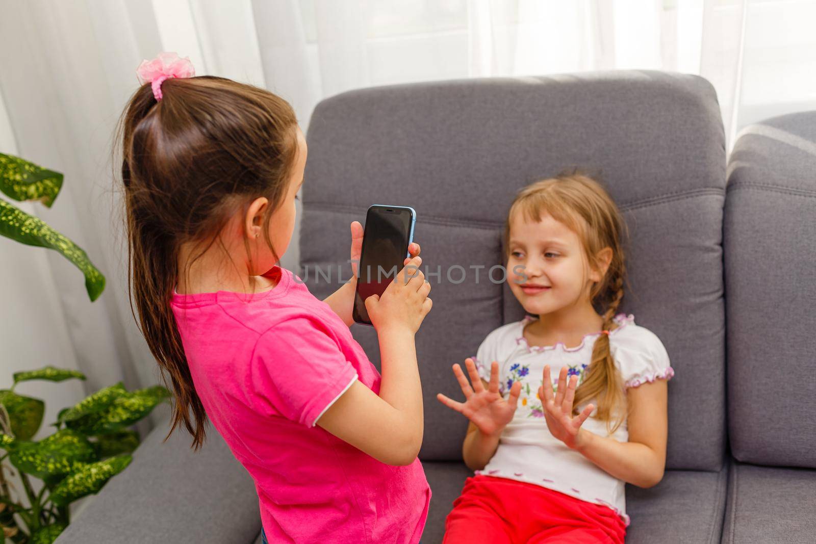 people, children, technology, friends and friendship concept - happy little girls sitting on sofa and taking selfie with smartphone at home