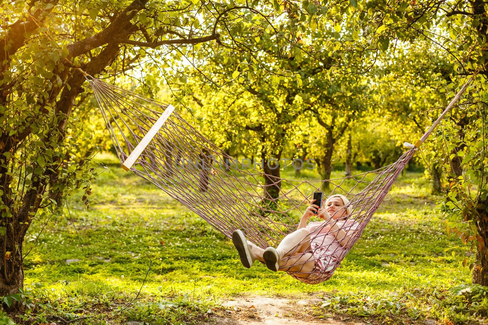 Smiling blonde woman relaxing on the hammock in garden, leisure time and summer holiday concept.