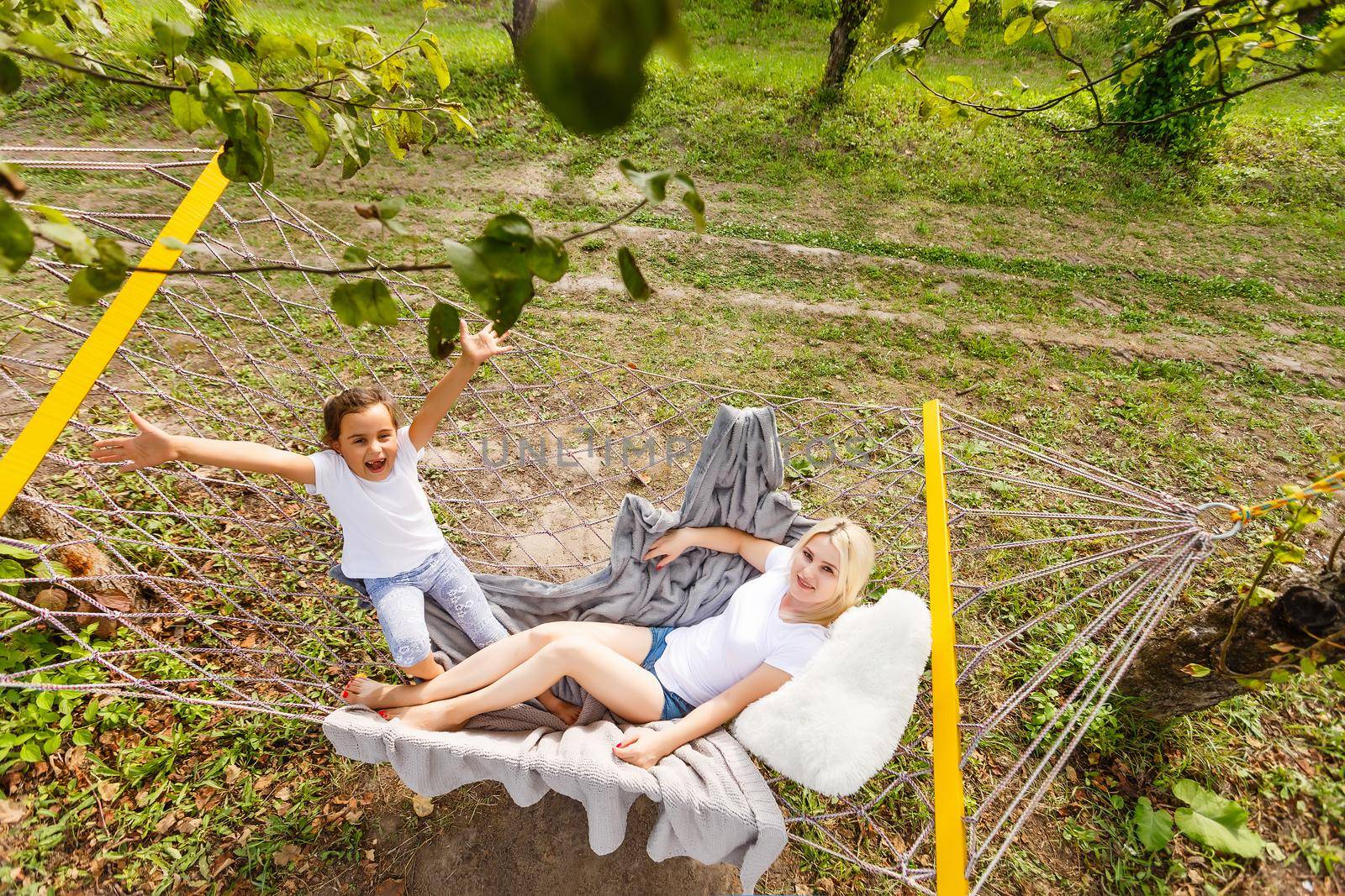 Wide view of a young mother and daughter relaxing together and smiling sitting in a hammock, hugging and lounging during a sunny summer day in a holiday home garden with grass and trees, lifestyle.