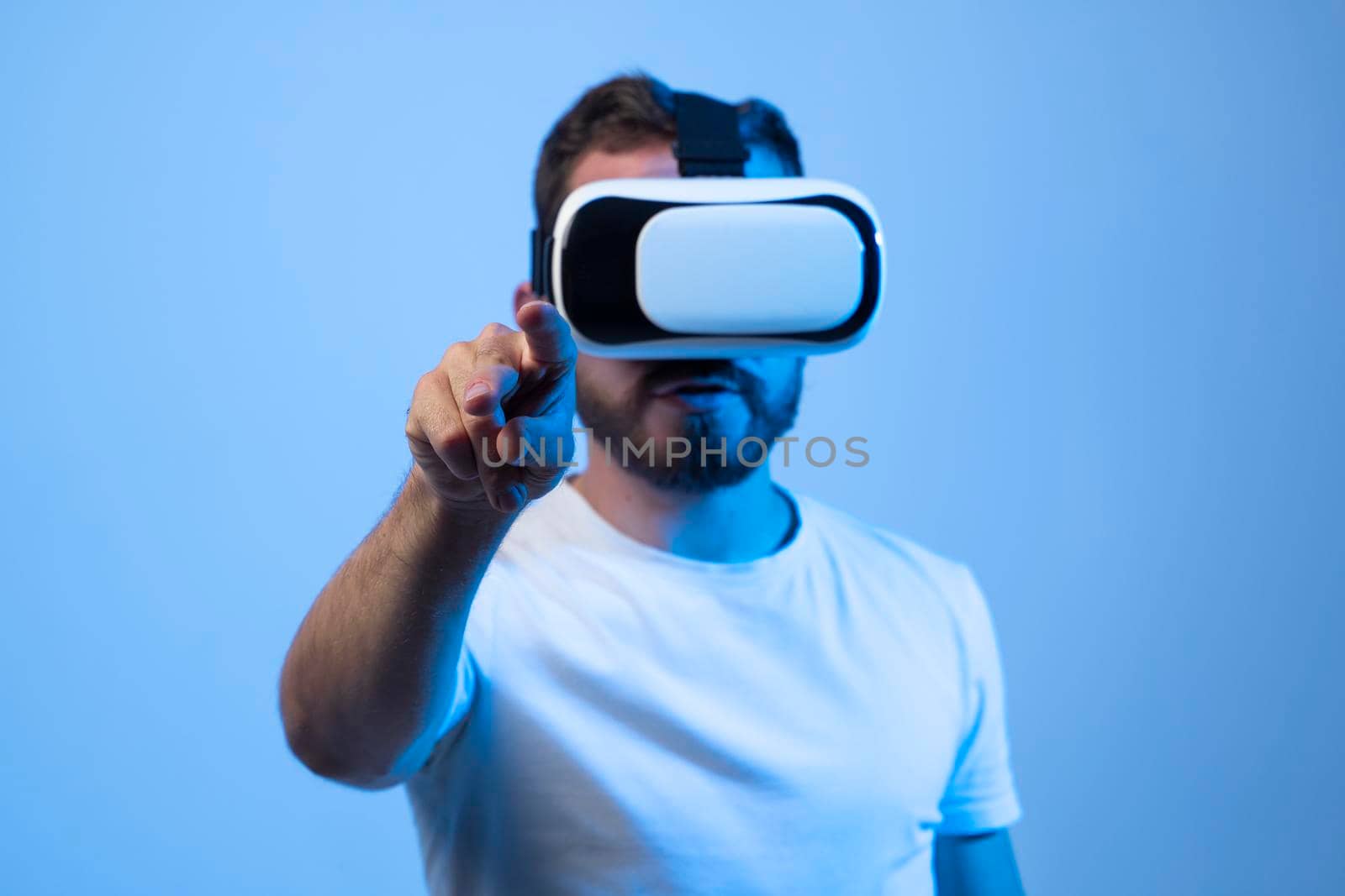 Bearded man student in white t-shirt uses vr glasses and try to touch something with finger, studio shot. Virtual gadgets for entertainment, work, free time and study
