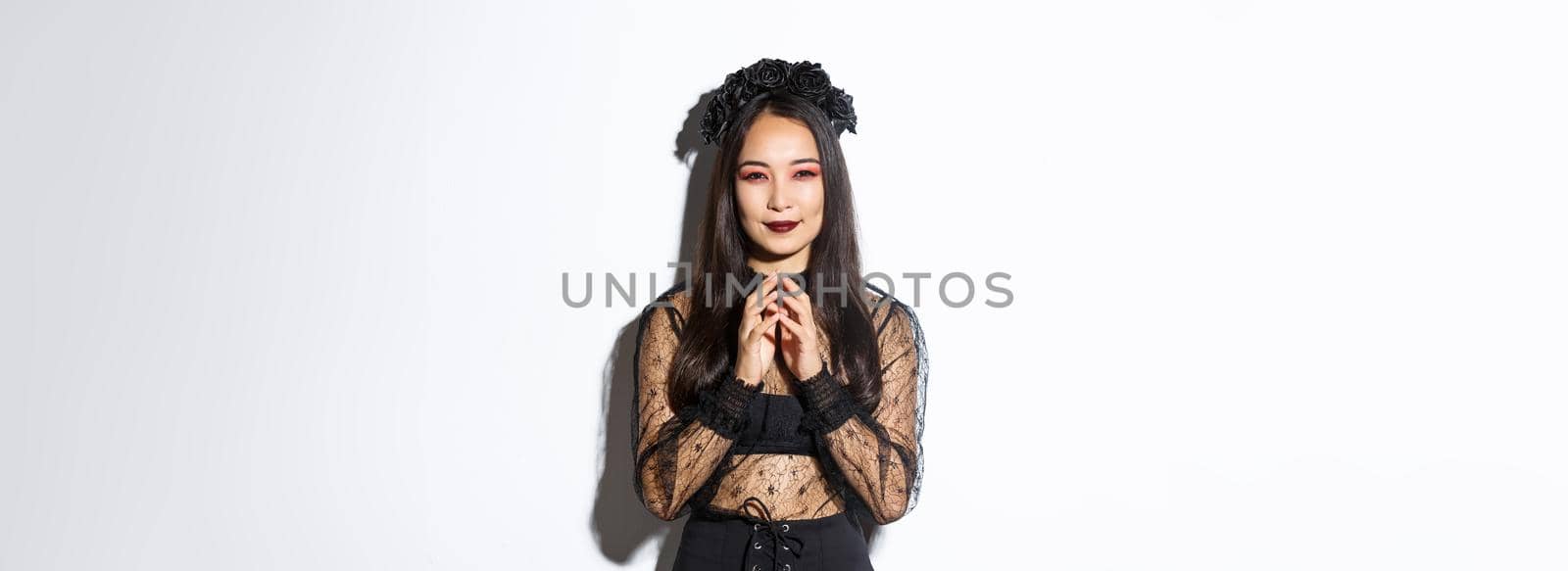 Devious young asian woman in gothic lace dress smiling pleased and steeple fingers. Witch prepare evil plan, smiling cunning and looking at camera. Halloween concept.