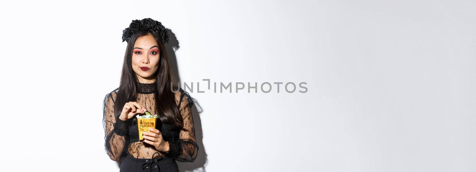 Confident smiling asian woman looking satisfied, holding sweets and wearing witch costume on halloween, standing over white background.