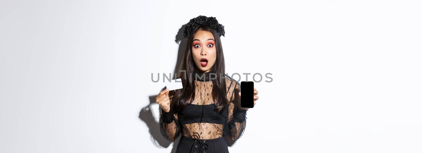 Amazed asian girl in witch costume showing credit card with mobile phone screen, open mouth surprised, celebrating halloween, white background.