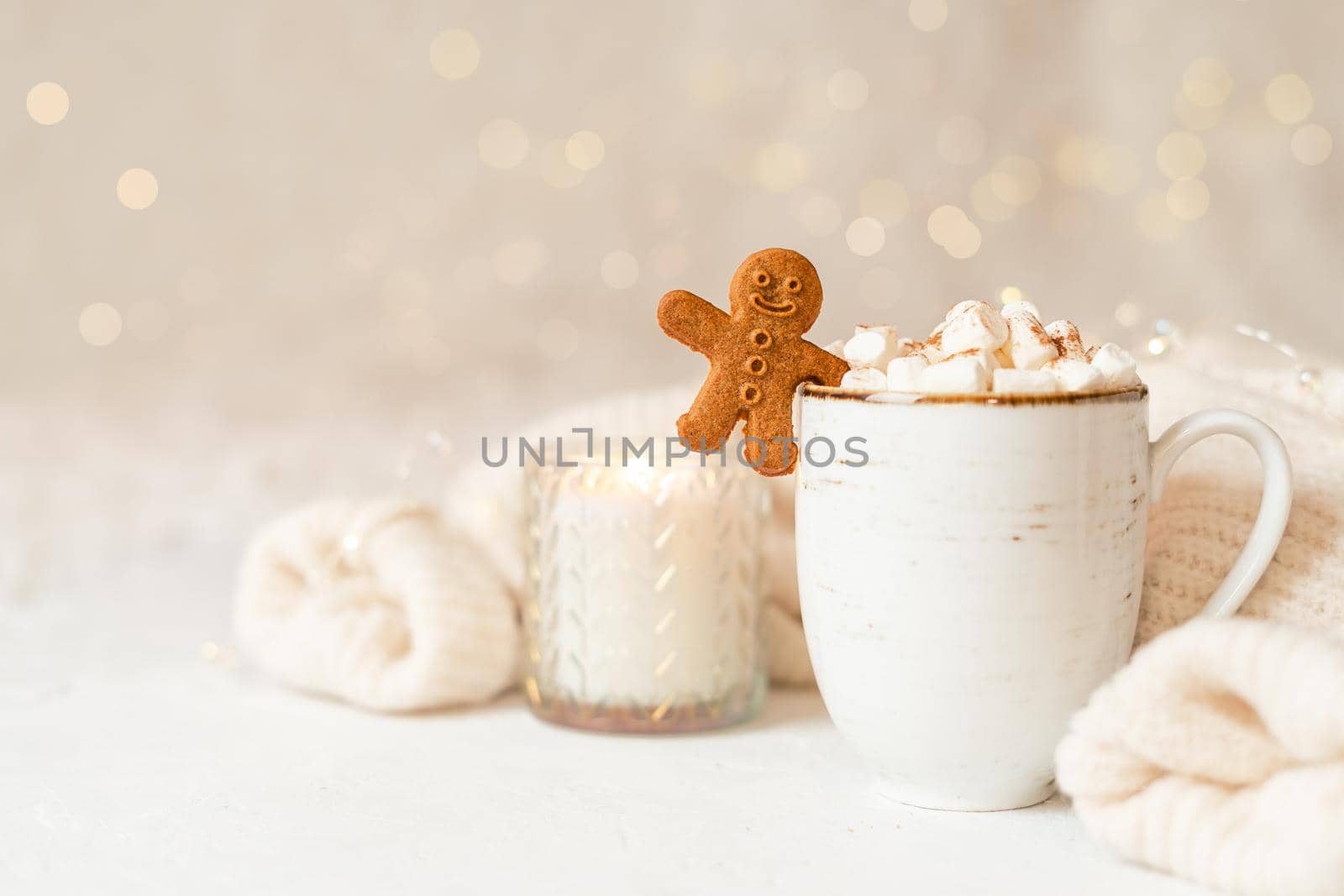 Gingerbread man cookie in cup of hot cocoa or coffee with marshmallow, candle, cinnamon and warm cozy sweater. Christmas lights background. Xmas holiday wooden star decorations.